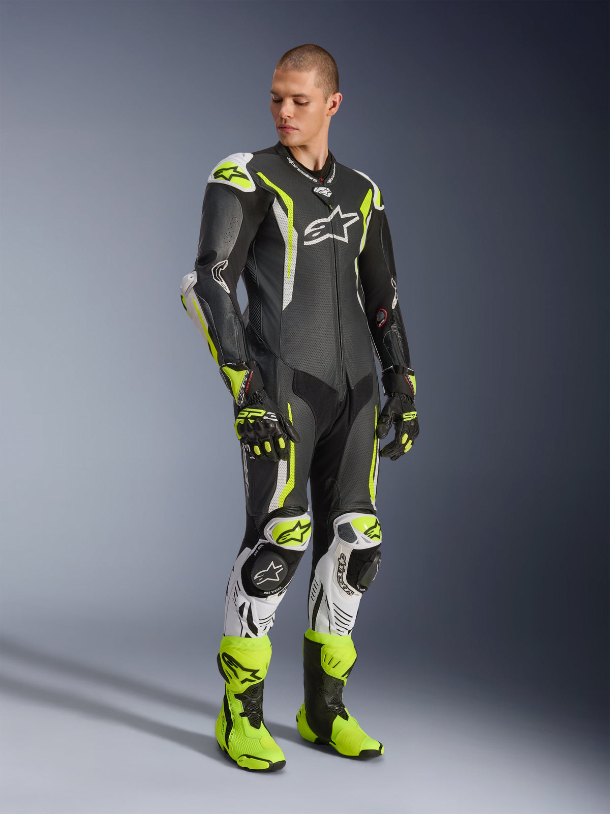 Alpinestars GP Tech V5 One Piece Leather Suit, czarno-biało-żółty fluo, prezentowany przez modela z dopasowanymi rękawicami i butami wyścigowymi, profesjonalny motocyklowy ubiór wyścigowy o właściwościach aerodynamicznych z ochronnymi ochraniaczami.