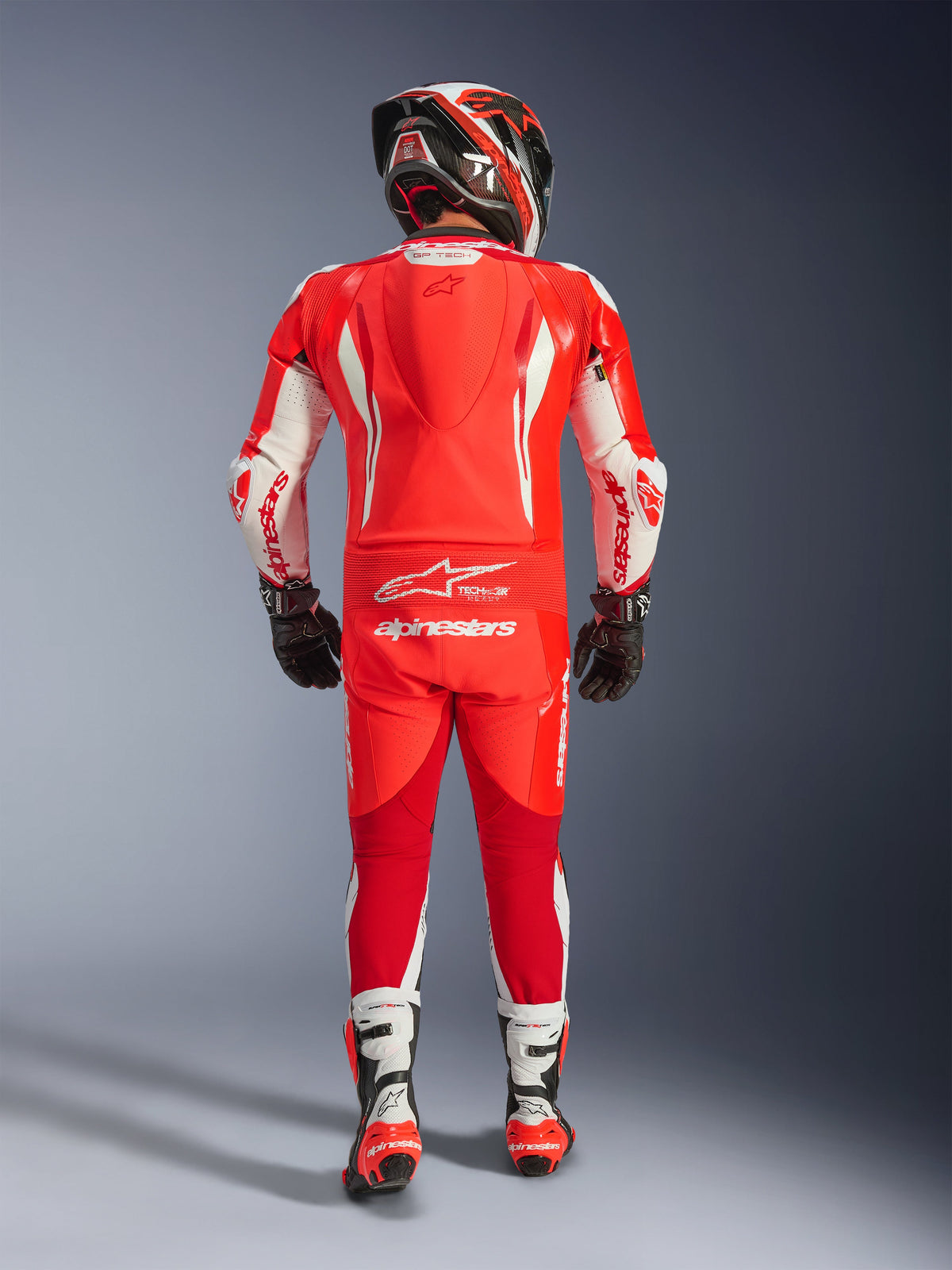 Widok z tyłu motocyklisty w jednoczęściowym kombinezonie skórzanym Alpinestars GP Tech V5 w kolorach Red Fluo, Bright Red i białym; wysokiej klasy kombinezon z aerodynamicznym garbem, białymi akcentami i logo Alpinestars, zestawiony z pasującym kaskiem, czarnymi rękawicami oraz biało-czerwonymi butami wyścigowymi.