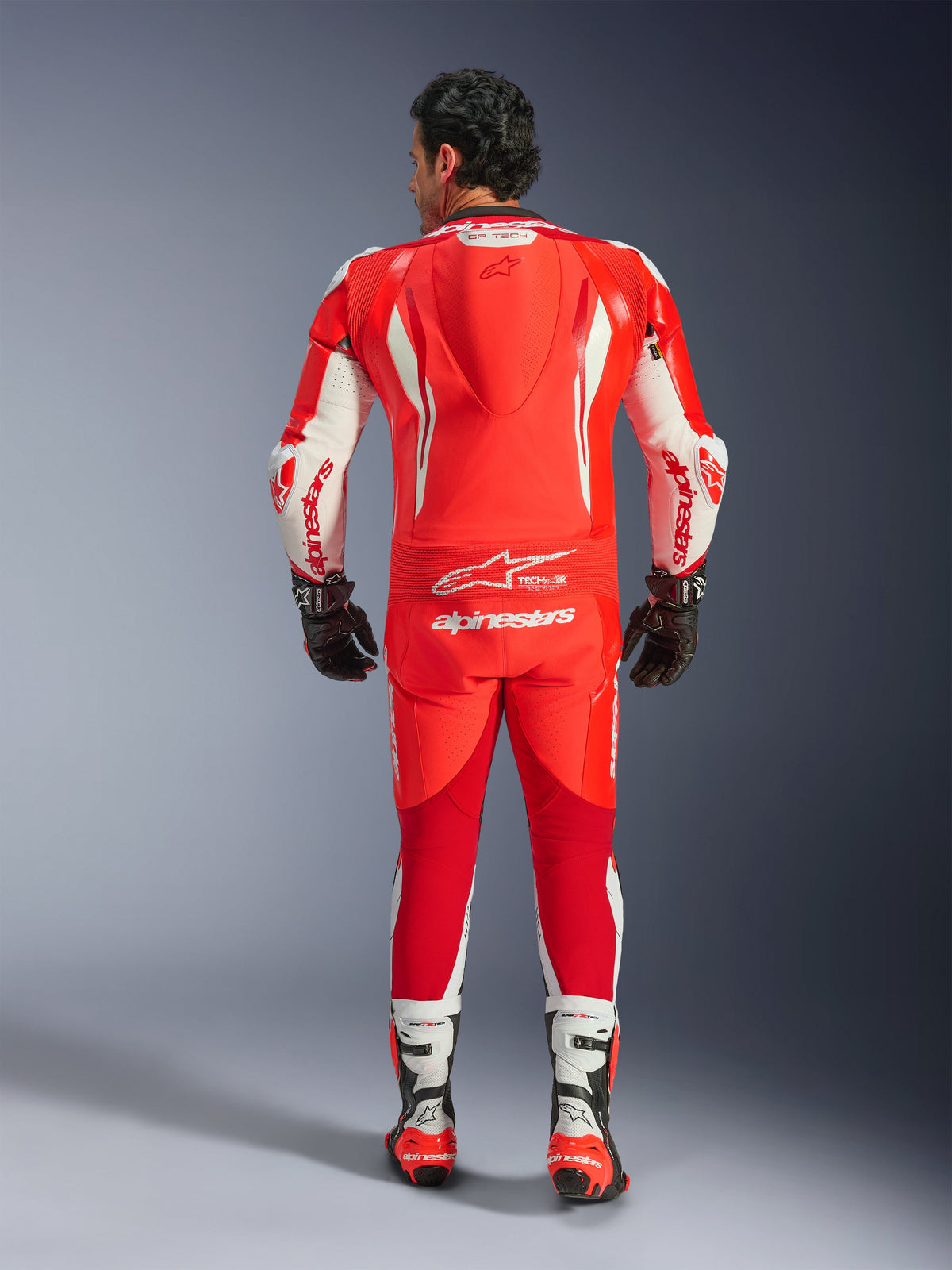 GP Tech V5 One Piece Leather Suit, wyścigowy kombinezon motocyklowy, Red Fluo Bright Red i biały, widok z tyłu pokazujący garb aerodynamiczny i białe logo Alpinestars, noszony z czarno-białymi rękawicami i butami wyścigowymi.