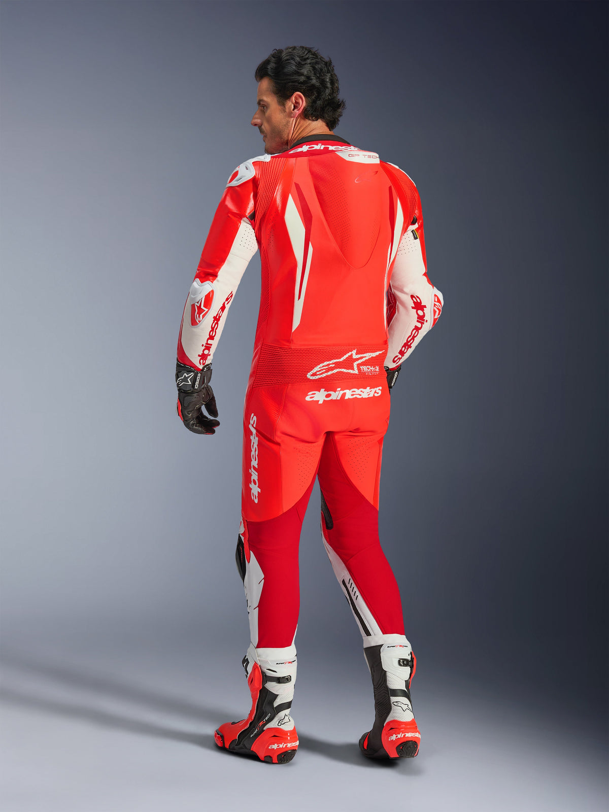 Alpinestars GP Tech V5 One Piece Leather Suit, jednoczęściowy kombinezon skórzany, fluorescencyjna czerwień, jasnoczerwony i biały, widok z tyłu ukazujący garb aerodynamiczny i logo Alpinestars, czarne rękawice i biało-czerwone buty wyścigowe, profesjonalna odzież motocyklowa