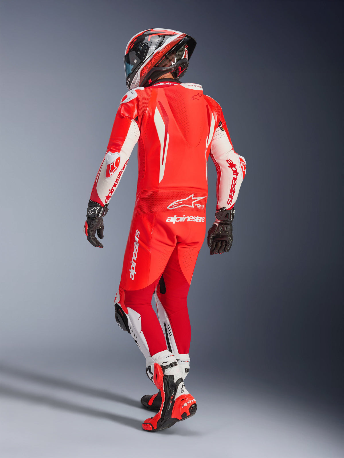 Alpinestars GP Tech V5 One Piece Leather Suit, kolor Red Fluo, jaskrawoczerwony i biały, widok z tyłu motocyklisty w pełnym stroju wyścigowym, aerodynamiczny garb i białe panele na rękawach i nogawkach, dopasowany kask i czerwone buty wyścigowe, profesjonalny design torowy