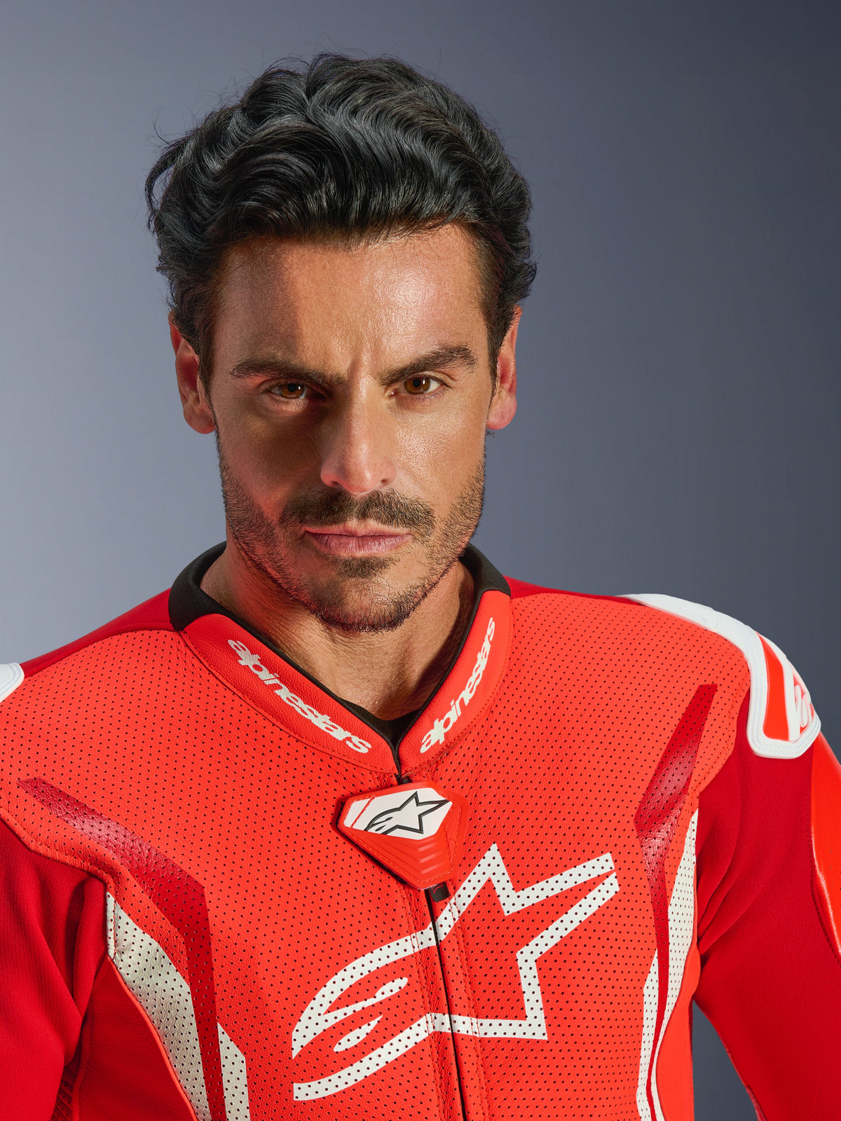 Alpinestars GP Tech V5 One Piece Leather Suit, motocyklowy kombinezon wyścigowy, jaskrawoczerwony i fluorescencyjny czerwony z białymi elementami, portret motocyklisty z bliska, perforowana skóra na klatce piersiowej z białym logo Alpinestars, wysokowydajny kołnierz wyścigowy i aerodynamiczny design.