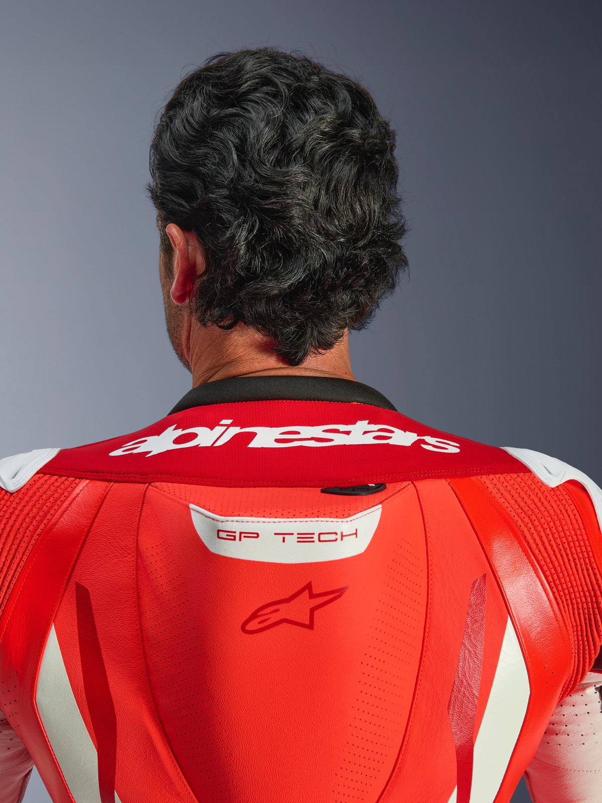 Zbliżenie na górną część pleców i kołnierz jednoczęściowego kombinezonu skórzanego GP Tech V5, w kolorach Red Fluo i Bright Red, z białym napisem Alpinestars na kołnierzu i logo GP TECH na garbie aerodynamicznym, noszonego przez motocyklistę.