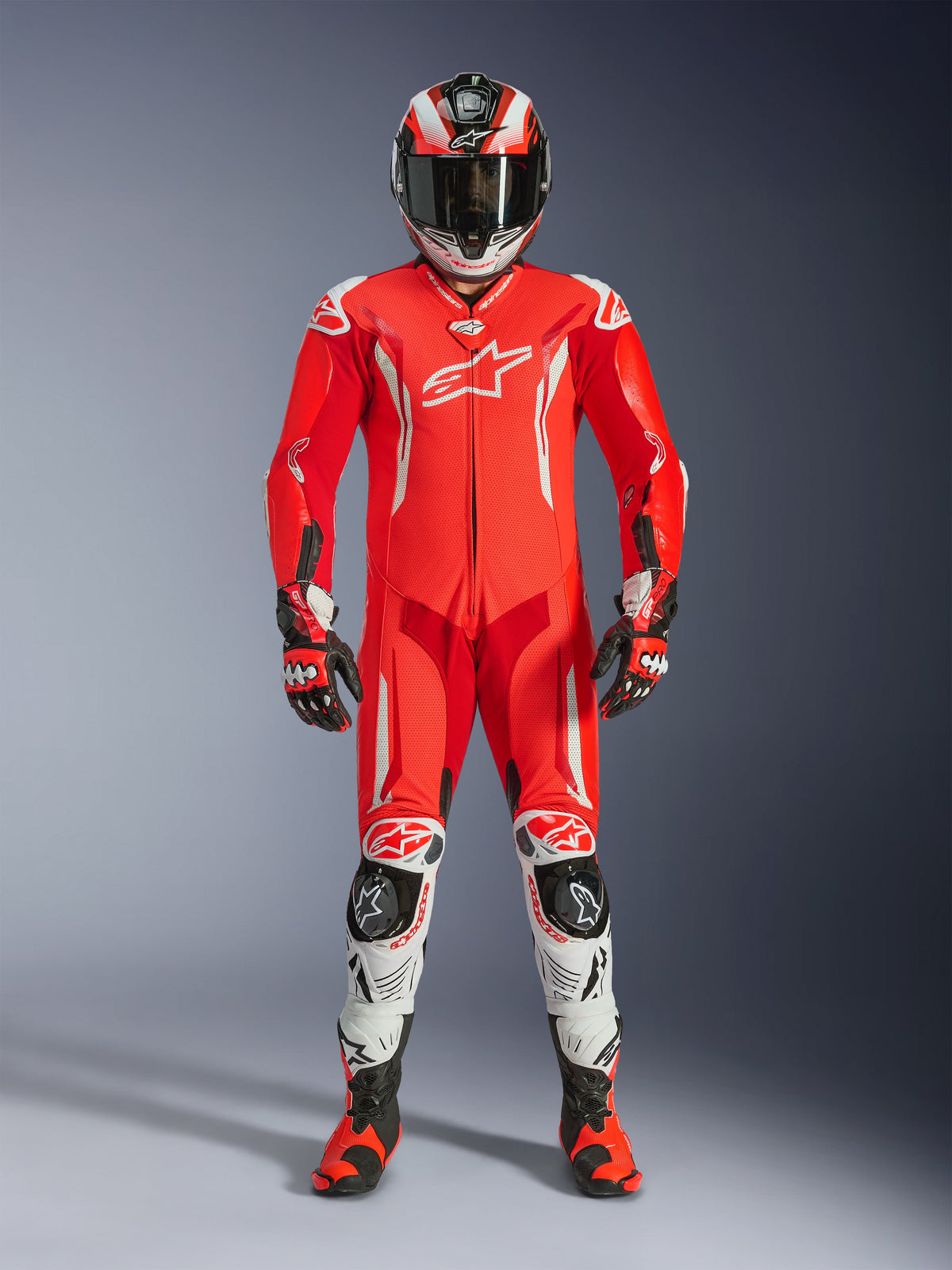 Motocyklista ubrany w jednoczęściowy skórzany kombinezon Alpinestars GP Tech V5 w kolorach Red Fluo, Bright Red i białym, w komplecie z pasującym kaskiem wyścigowym, rękawicami i butami. Profesjonalny kombinezon torowy o aerodynamicznej sylwetce, z białymi logotypami Alpinestars i zintegrowanymi sliderami ochronnymi.