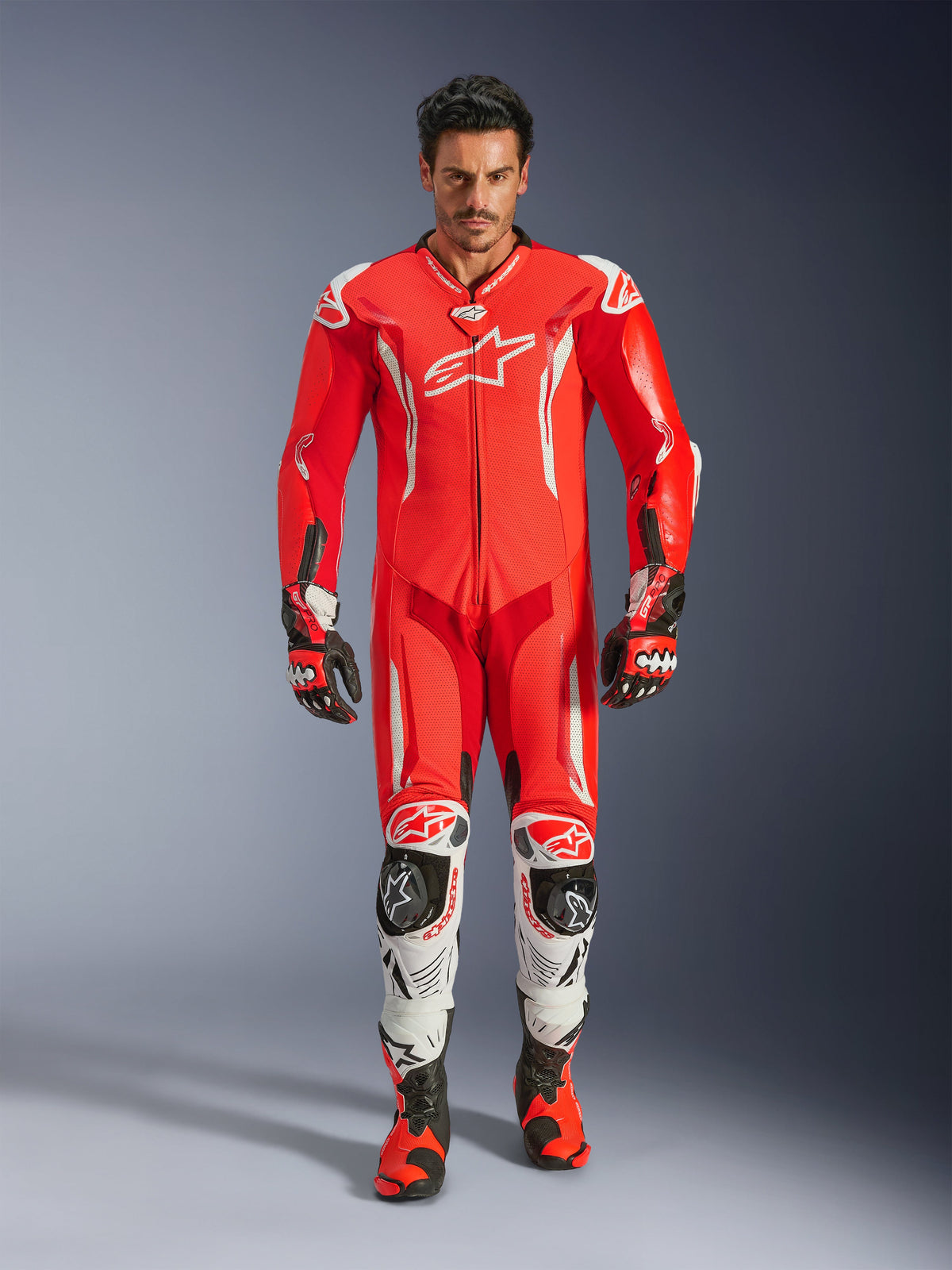 Alpinestars GP Tech V5, jednoczęściowy kombinezon skórzany, profesjonalny kombinezon wyścigowy w kolorach Red Fluo i Bright Red z białymi akcentami, noszony przez motocyklistę z dopasowanymi butami i rękawicami, aerodynamiczny garb, skórzana konstrukcja premium i zaawansowane ochraniacze.