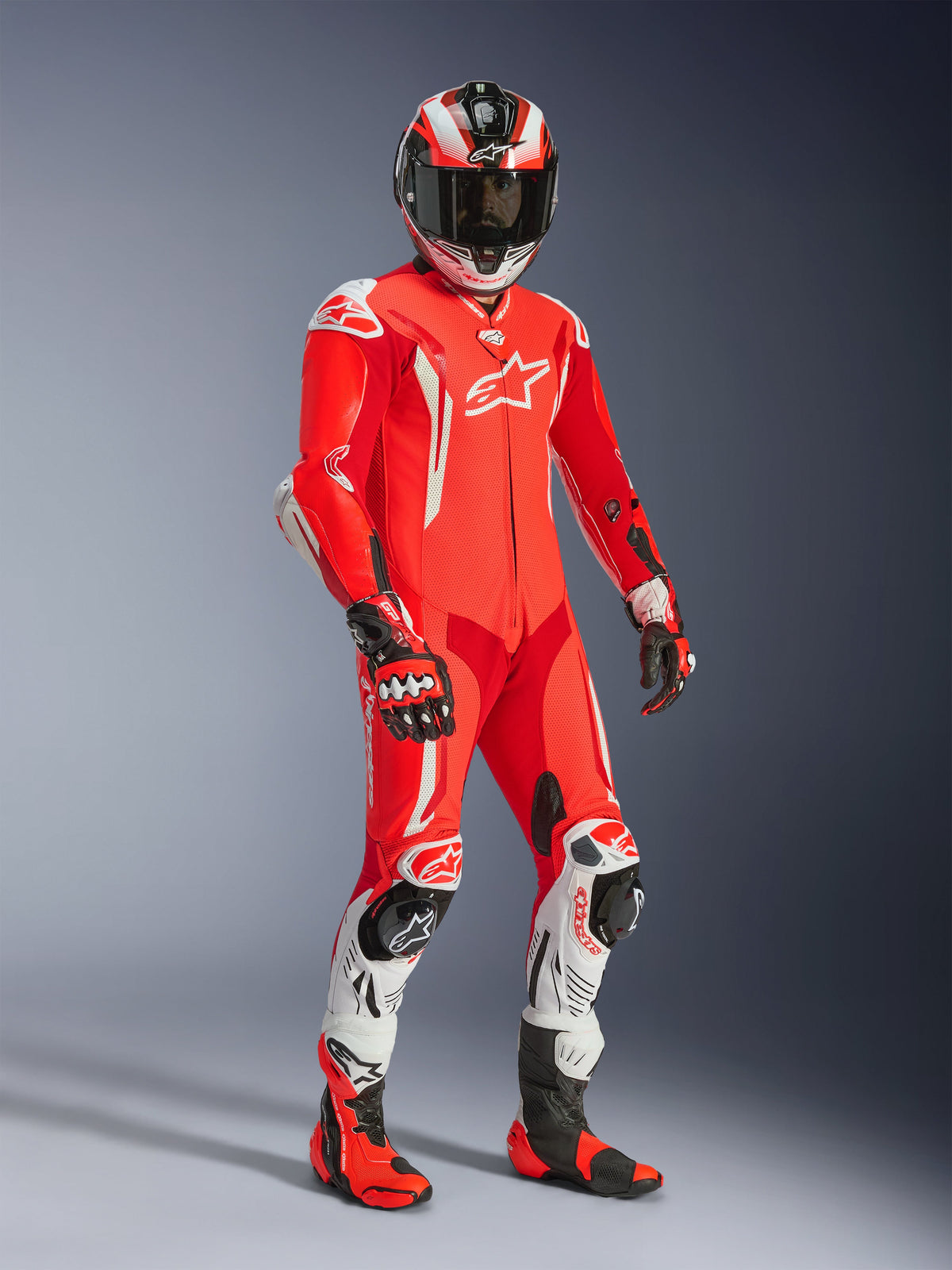 Motocyklista w jednoczęściowym kombinezonie skórzanym Alpinestars GP Tech V5, fluorescencyjna czerwień i jasna czerwień z białymi akcentami, z dopasowanym kaskiem, rękawicami i butami wyścigowymi, profesjonalny sprzęt torowy z perforowaną skórą i sliderami ochronnymi