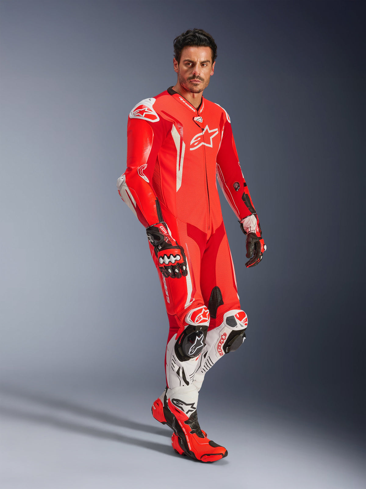 Mężczyzna w jednoczęściowym kombinezonie skórzanym Alpinestars GP Tech V5, kolor fluorescencyjna czerwień, jaskrawa czerwień i biały, dopasowane rękawice i buty wyścigowe, profesjonalna odzież motocyklowa z garbem aerodynamicznym i zintegrowaną ochroną