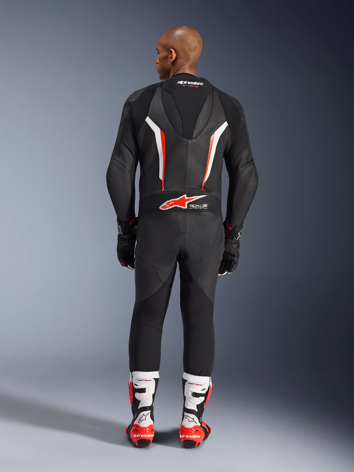 Rear view of a model wearing the Alpinestars GP Force V2 1-Piece Leather Suit in Black White Red Fluo, highlighting the aerodynamic speed hump and Tech-Air Ready branding, paired with white and red professional racing boots and black gloves.