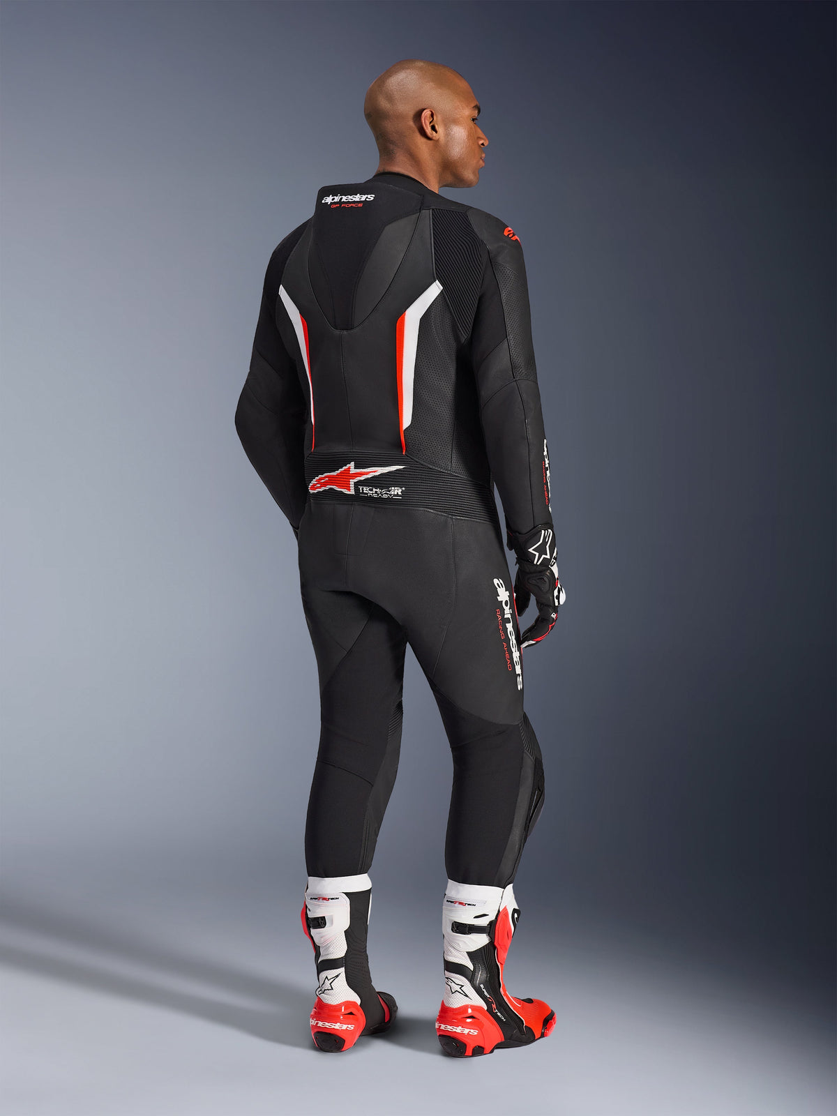 Rider wearing the GP Force V2 Leather Suit 1-Piece, Black with White and Red Fluo accents, seen from the back showing the aerodynamic speed hump and Alpinestars branding, paired with matching racing gloves and white and red performance boots, professional motorcycle racing gear