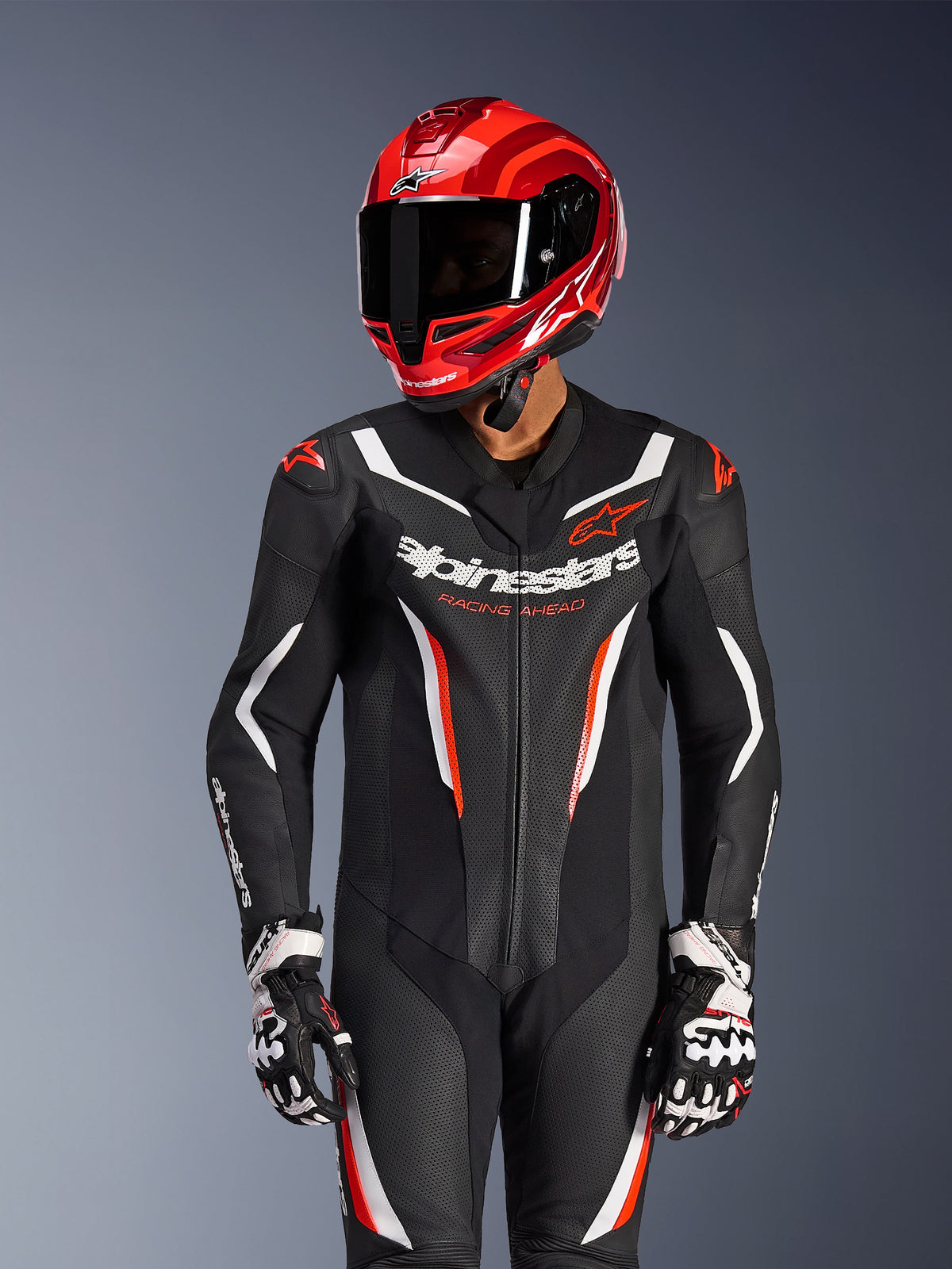 Motorcycle rider wearing Alpinestars GP Force V2 1-Piece Leather Suit in Black White Red Fluo, coordinated red racing helmet and gloves, features perforated leather panels and bold branding, high-performance racing gear
