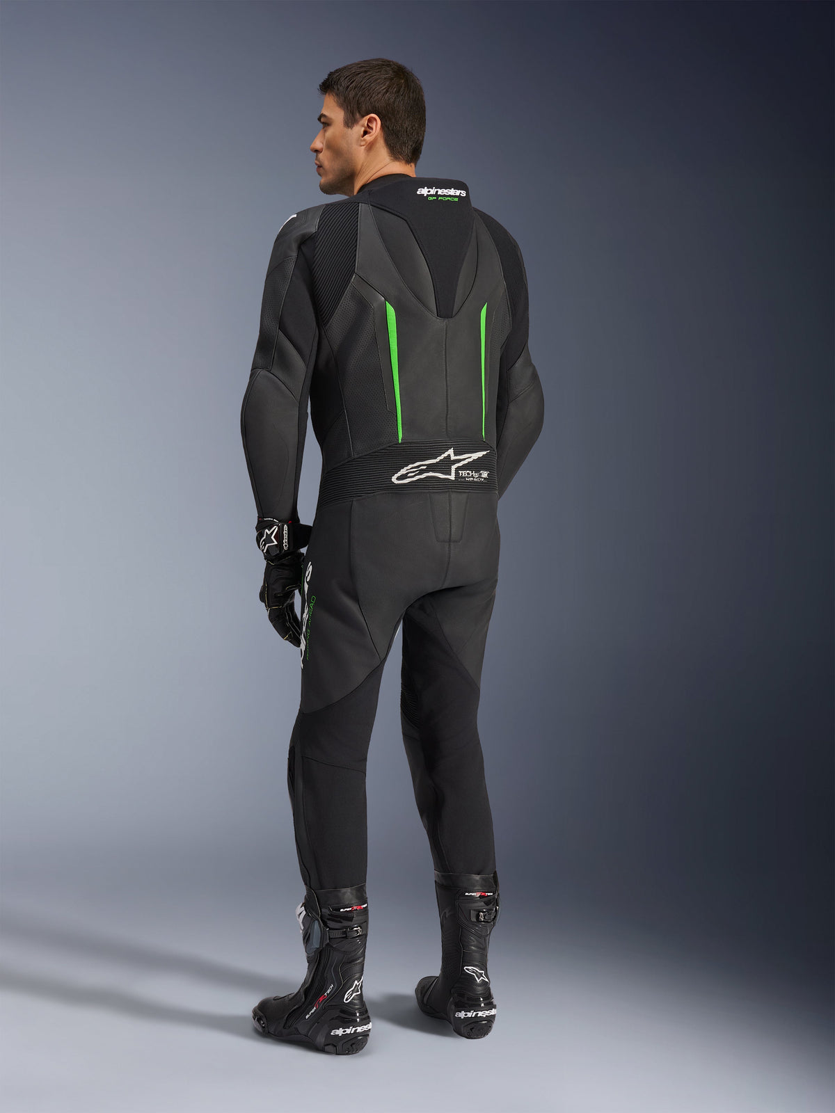 Alpinestars GP Force V2 Leather Suit, 1-Piece Motorcycle Suit, Black with Green accents, back view showing the aerodynamic speed hump and white logo on the accordion stretch panel, paired with black racing boots and gloves.