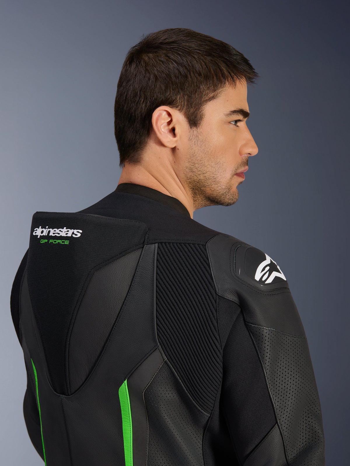 Close-up of the upper back and shoulder of a male model wearing the Alpinestars GP Force V2 Leather Suit in Black and Green, featuring the aerodynamic speed hump with green 'GP FORCE' branding, a white Astars logo on the shoulder, and accordion stretch panels.