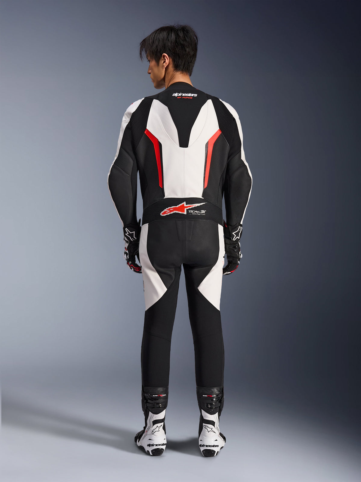 Alpinestars GP Force V2 Leather Suit 1-Piece, Motorcycle Race Suit, White Black and Red Fluo, back view of model wearing the suit with racing boots and gloves, featuring an aerodynamic speed hump, accordion stretch panels, and Tech-Air Ready branding