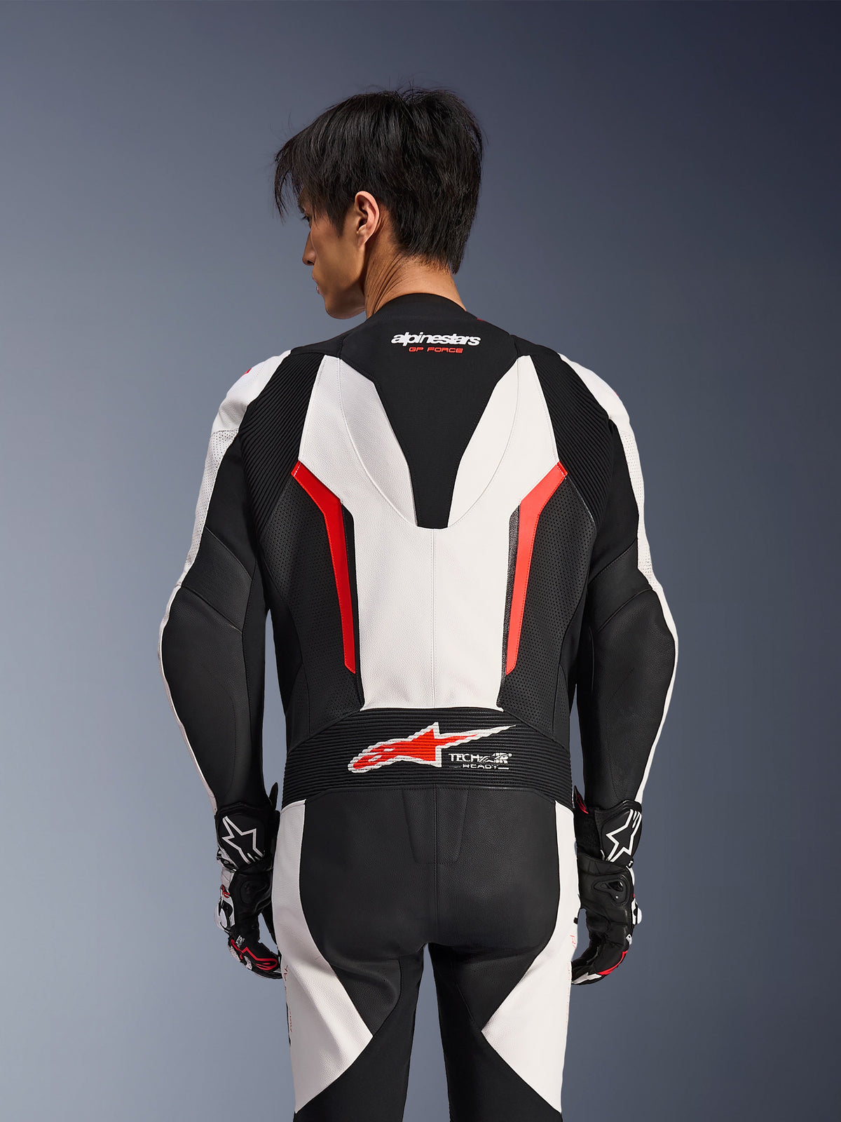 Alpinestars GP Force V2 Leather Suit 1Piece, White Black and Red Fluo, back view on male model, featuring aerodynamic speed hump, perforated leather panels, and Tech-Air Ready logo on the lower back, professional racing gear with black and white gloves