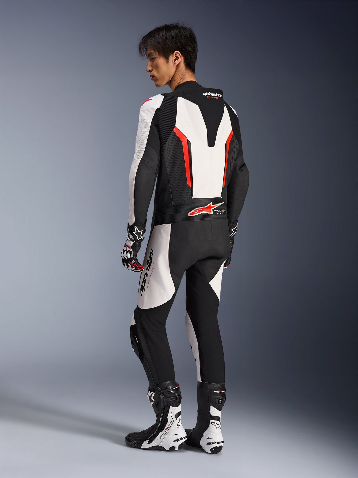 Motorcycle rider wearing the Alpinestars GP Force V2 Leather Suit - 1Piece in White Black and Red Fluo, rear-side view showing the aerodynamic speed hump and perforated leather panels, paired with matching racing boots and gloves, designed for track and performance riding.