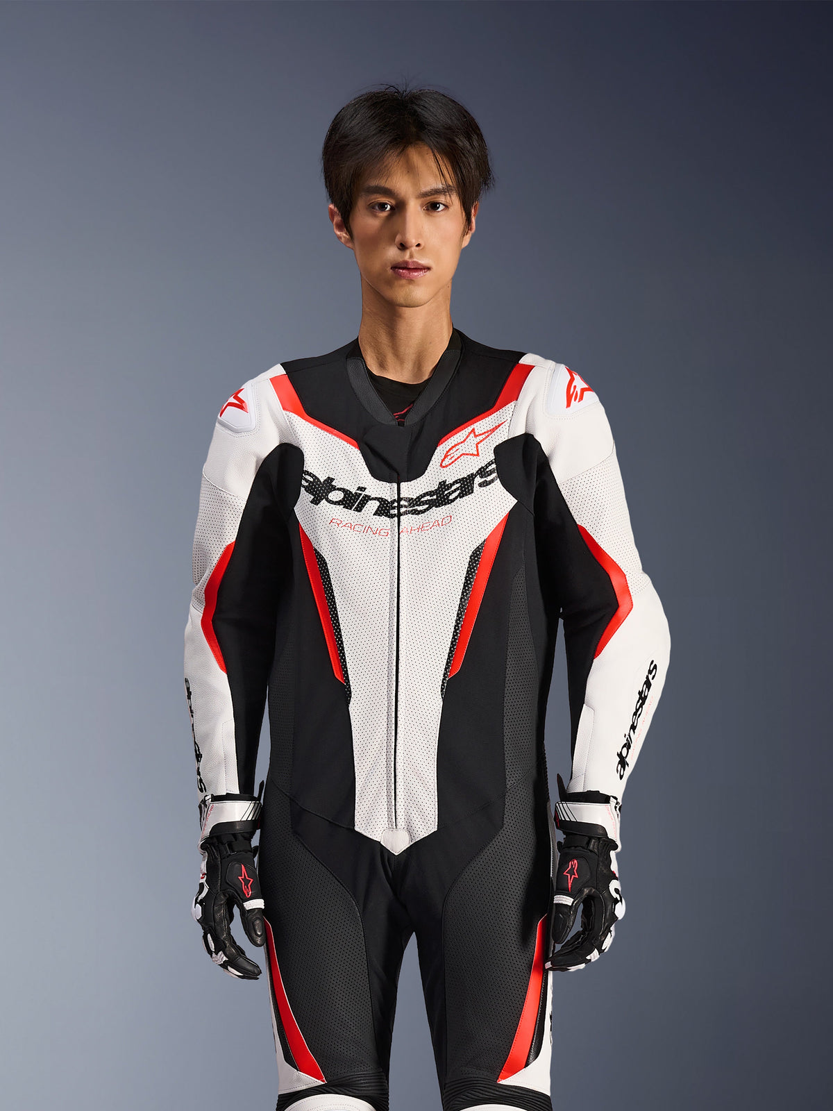 Alpinestars GP Force V2 Leather Suit - 1Piece, White Black and Red Fluo, worn by a male model, featuring perforated leather panels and bold Alpinestars branding on the chest, coordinated with black and white racing gloves, designed for track and performance riding