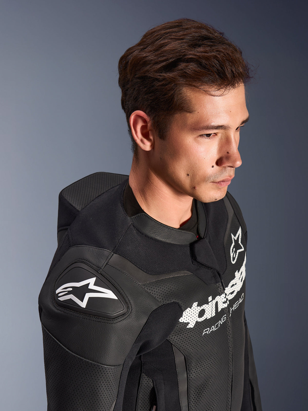Alpinestars GP Force V2 Leather Suit, 2-Piece, Black, worn by a model, featuring the shoulder with white Astar logo, chest branding, perforated leather panels, and aerodynamic speed hump, high-performance motorcycle racing gear