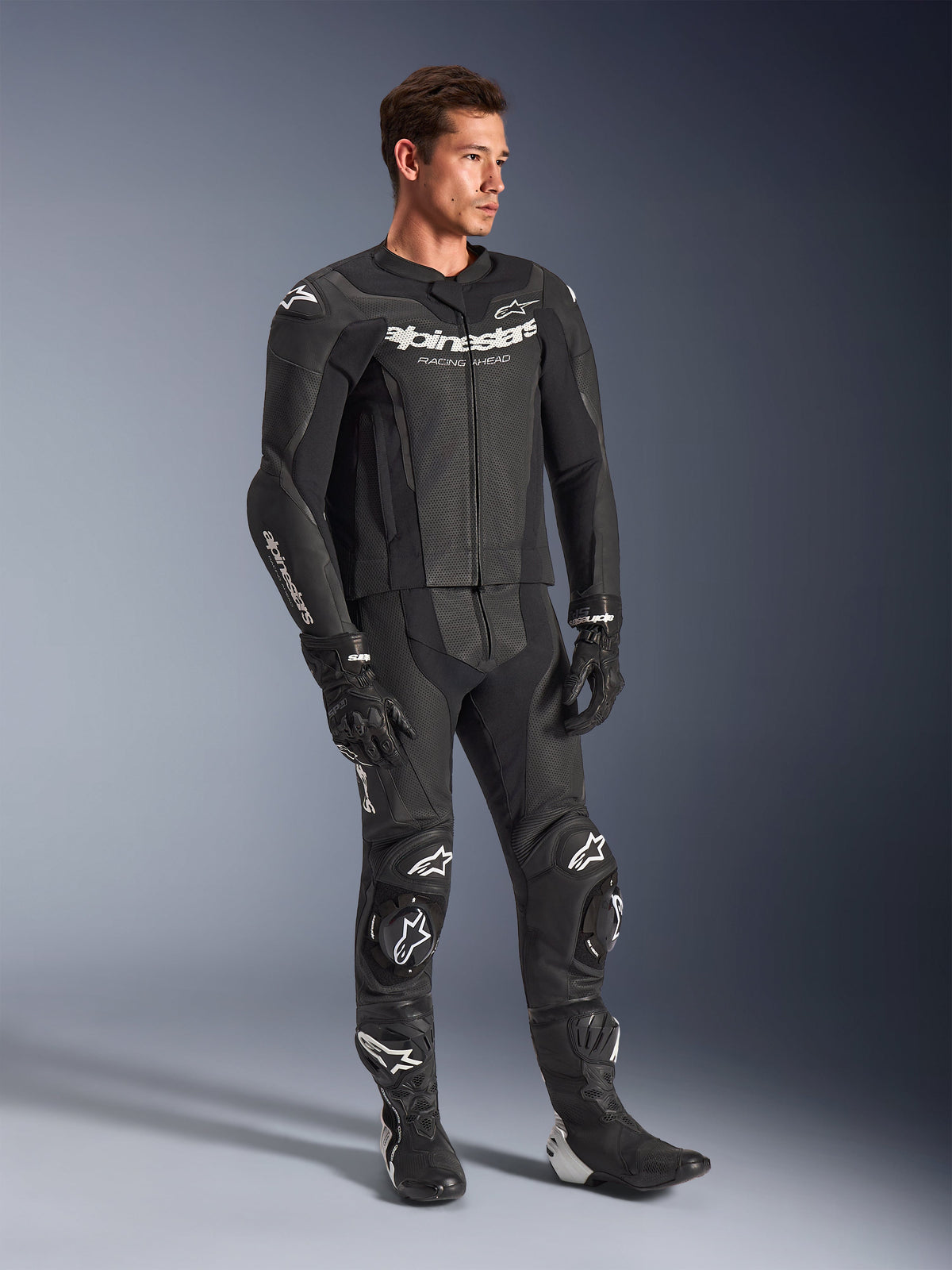 Alpinestars GP Force V2 2-Piece Leather Suit, Black, worn by a male model with matching black racing gloves and boots, featuring white Alpinestars branding, perforated leather panels for ventilation, and integrated knee sliders, professional motorcycle racing gear