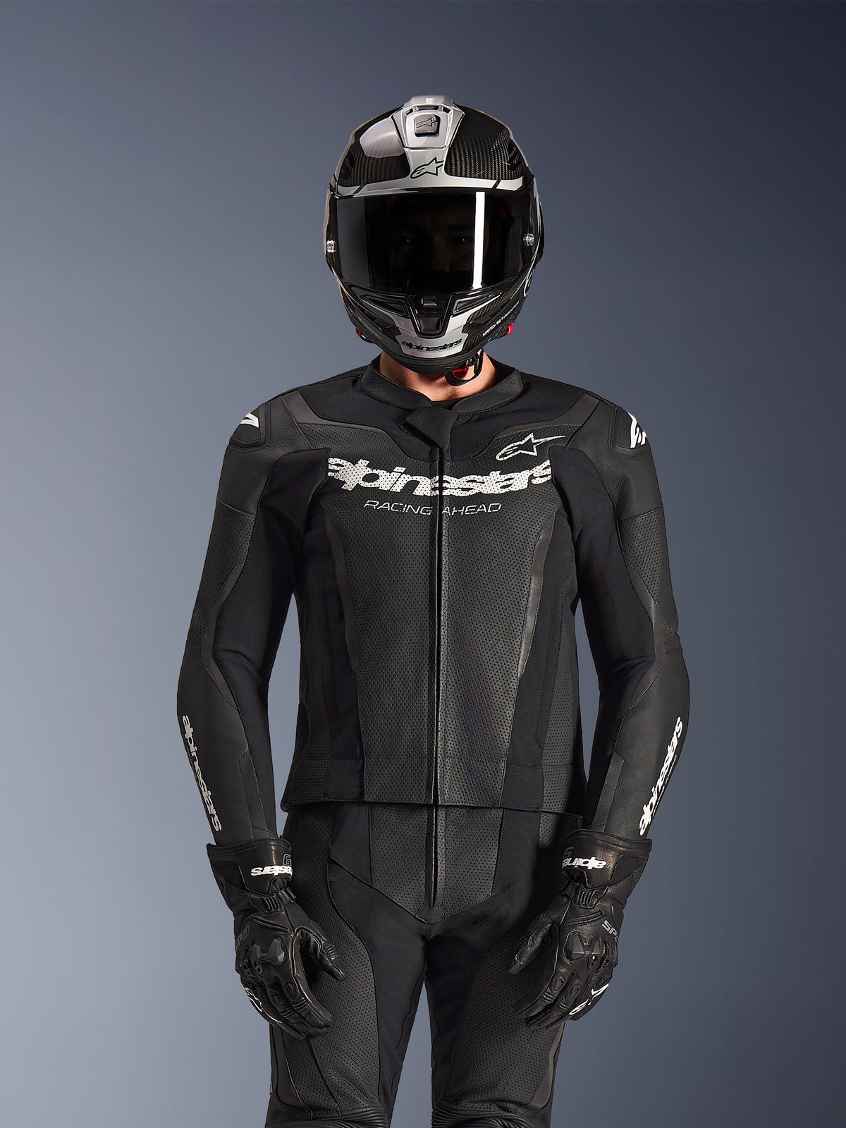 Rider wearing Alpinestars GP Force V2 2-Piece Leather Suit in Black, perforated leather construction with white 'Racing Ahead' chest logo, silver and carbon fiber full-face helmet, black gloves, professional sport and track riding gear