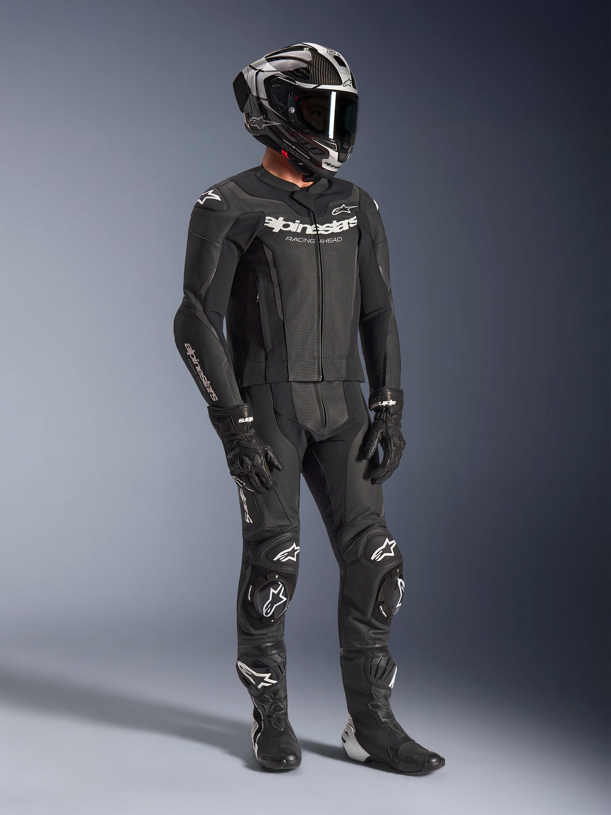 Alpinestars GP Force V2 2-Piece Leather Suit, black motorcycle racing suit, worn with a full-face helmet, gloves, and boots, featuring perforated leather construction, aerodynamic speed hump, and white Alpinestars logos on the chest, arms, and knees.