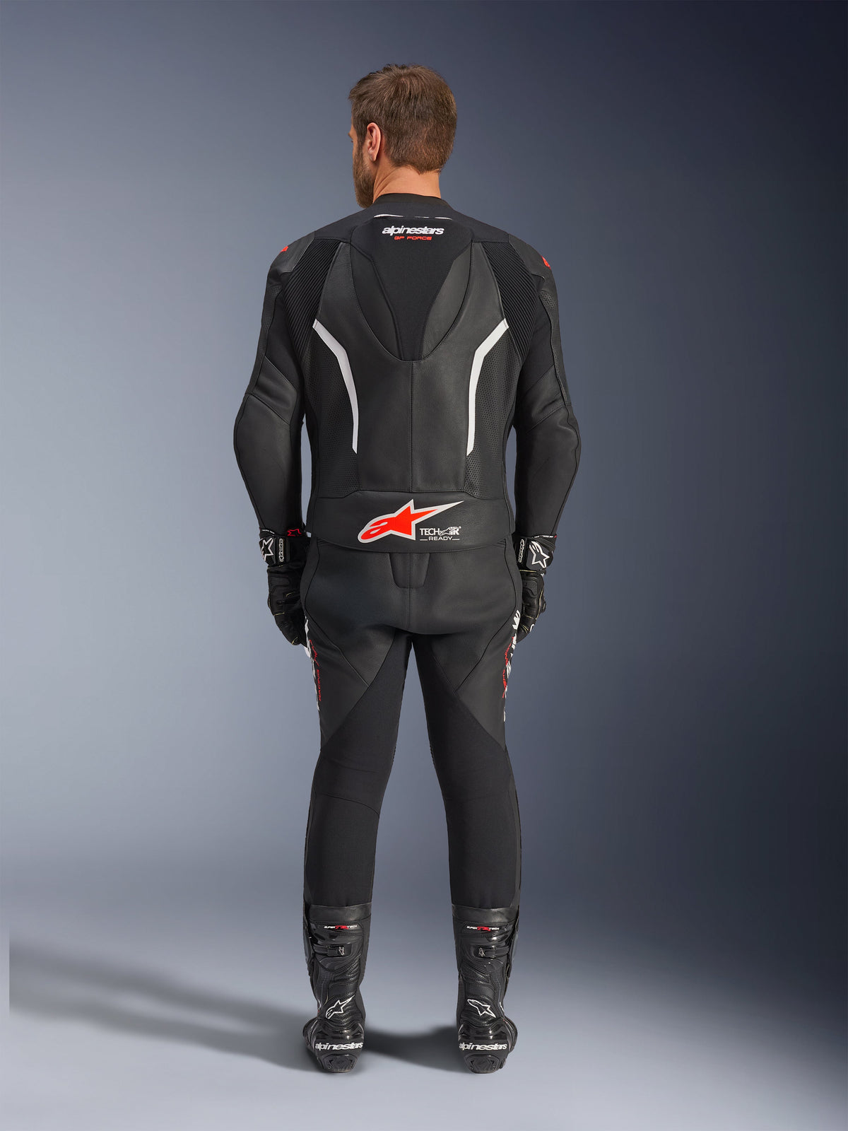 Rear view of a rider wearing the Alpinestars GP Force V2 2-Piece Leather Suit, Black with white and red accents, featuring prominent Alpinestars and Tech-Air Ready branding on the back, styled with matching black racing gloves and boots.