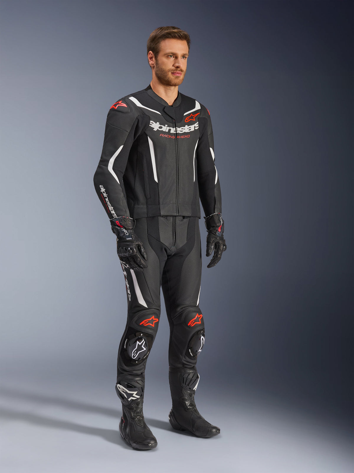 Alpinestars GP Force V2 2-Piece Leather Suit, Black White and Red Fluo, worn by a model with black racing boots and gloves, featuring perforated leather panels, white aerodynamic accents, and red fluo logos on shoulders and knees, professional motorcycle racing gear
