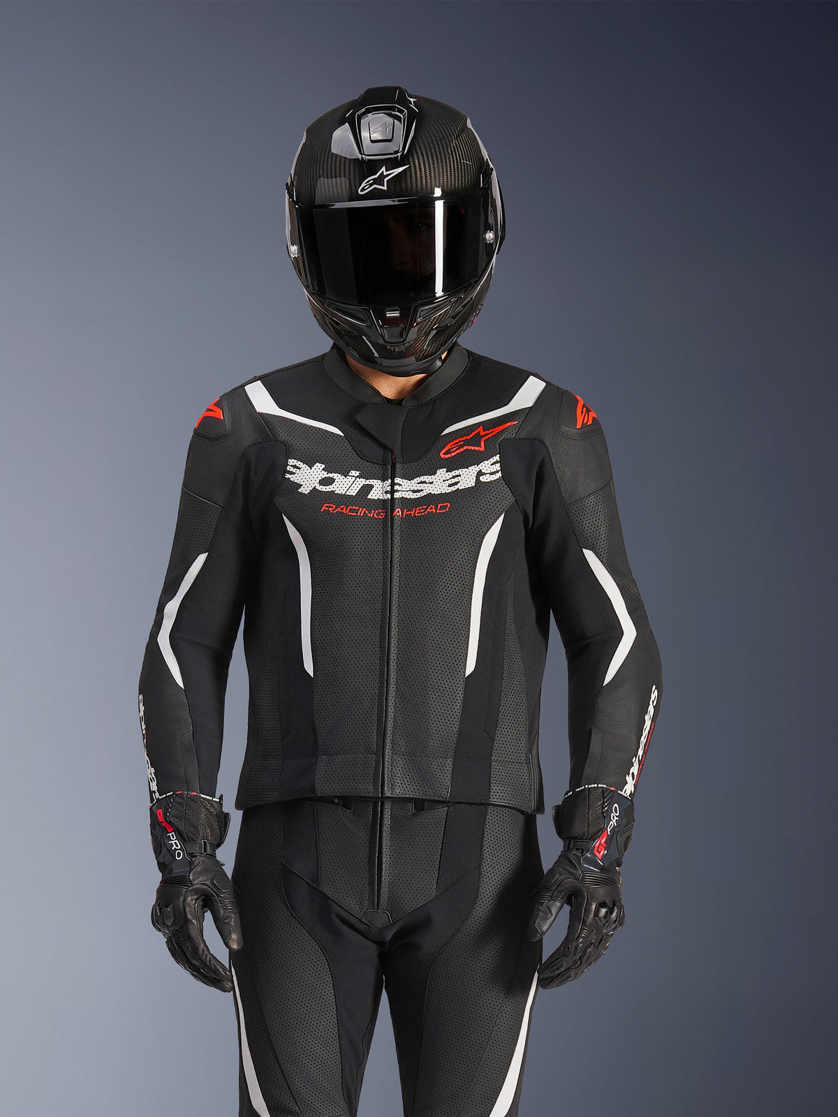Alpinestars GP Force V2 2-Piece Leather Suit, Black White and Red Fluo, worn by a rider with a carbon fiber full-face helmet and black racing gloves, featuring perforated leather panels and Alpinestars branding on the chest.