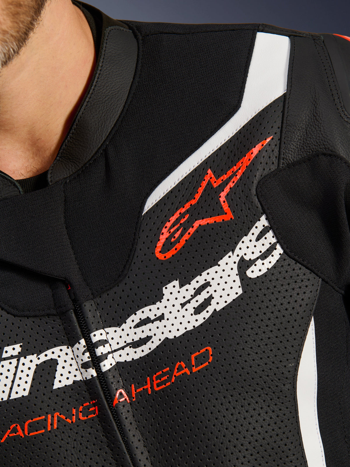 Alpinestars GP Force V2 Leather Suit 2-Piece, close-up of the chest, black perforated leather with white and red accents, featuring the red Alpinestars logo and 'RACING AHEAD' text, premium motorcycle racing apparel detail