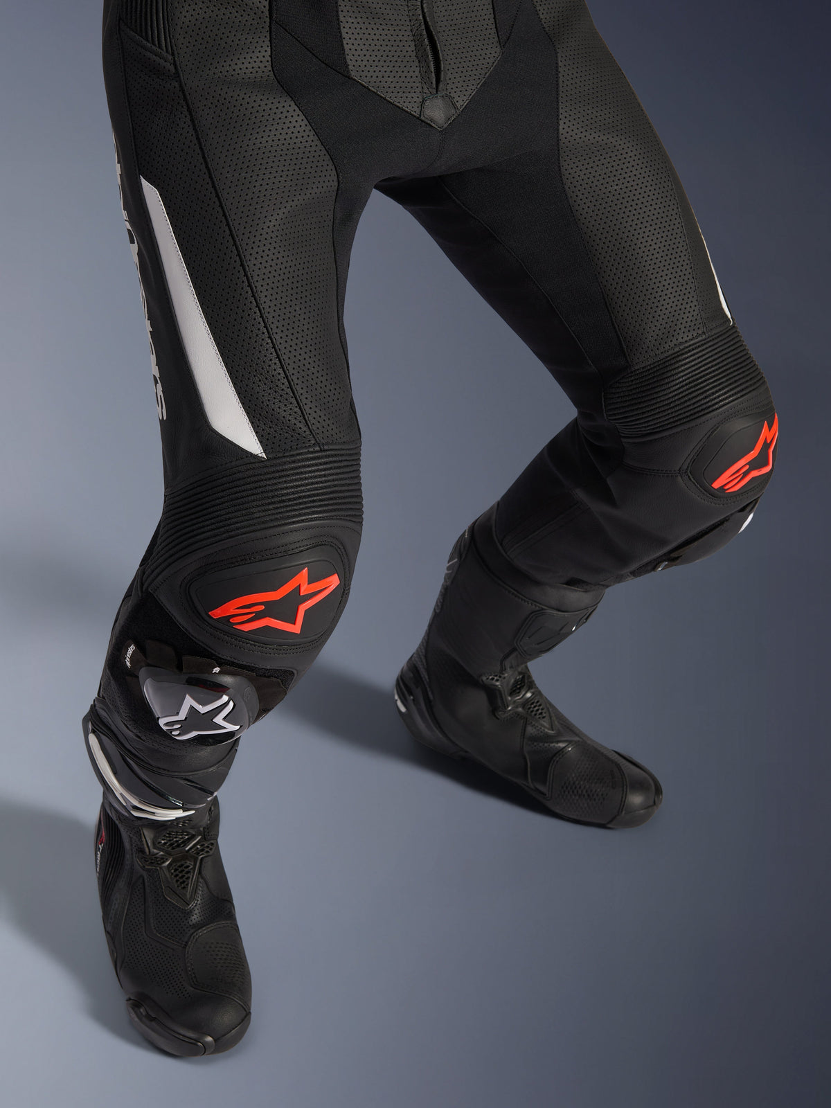 Alpinestars GP Force V2 2-Piece Leather Suit, motorcycle racing pants in black with red and white accents, featuring perforated leather, accordion stretch panels above the knees with red star logos, worn with black motorcycle boots, high-performance racing gear
