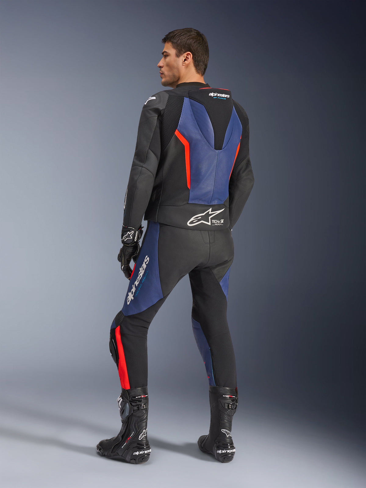 GP Force V2 Leather Suit 2-Piece, Black Blue and Red Fluo, rear profile view on a model, featuring aerodynamic hump, perforated leather, and Tech-Air Ready branding, worn with black Alpinestars racing boots and gloves
