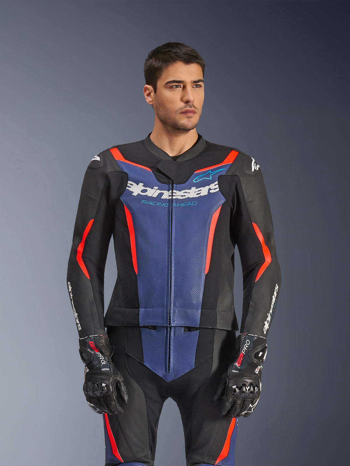 Male model wearing Alpinestars GP Force V2 Leather Suit 2-Piece, Black Blue Red Fluo, featuring bold blue chest panel and red accents, worn with GP Pro gloves, professional leather motorcycle racing gear with perforated texture