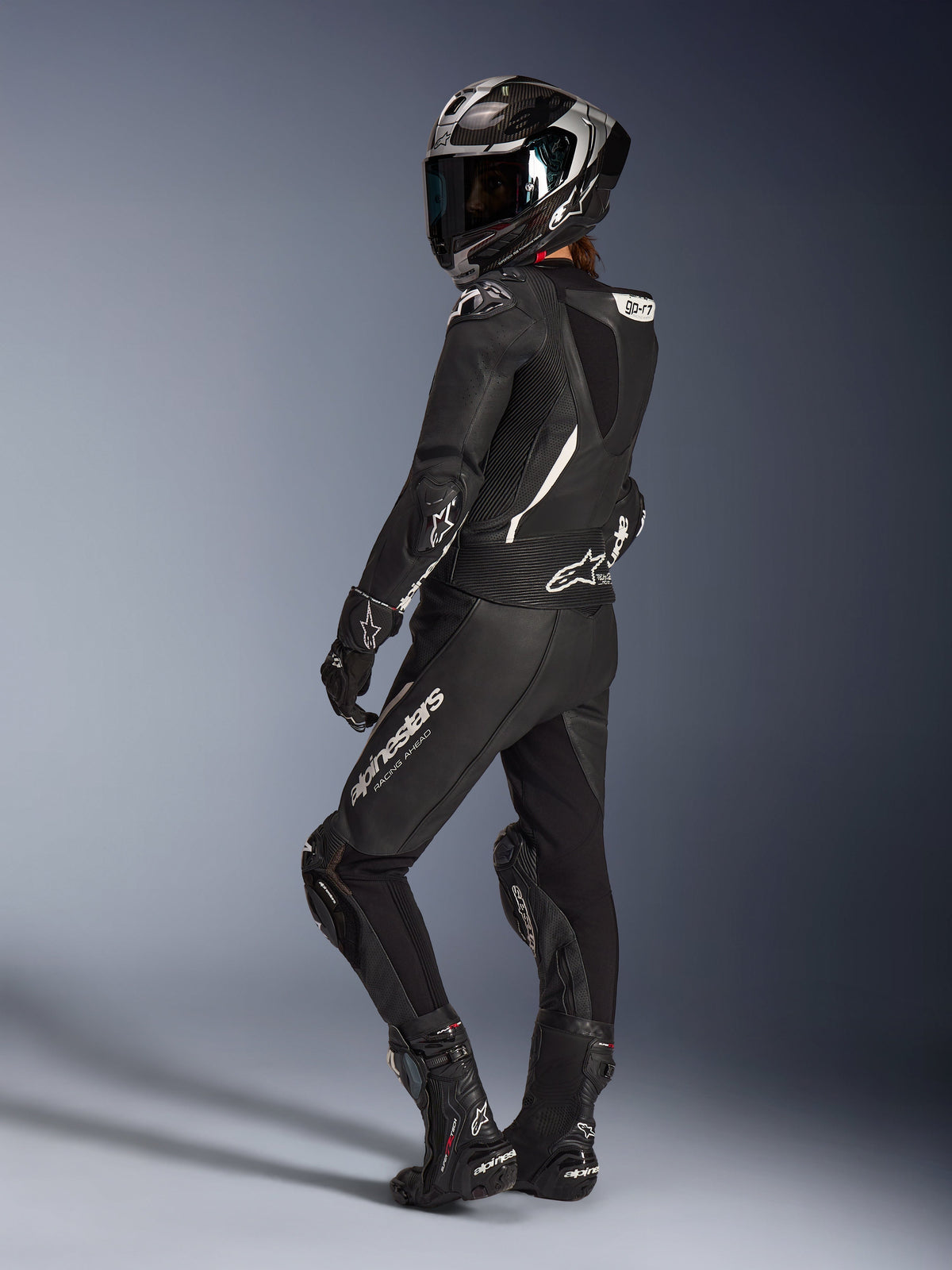 Motocyklistka w czarnym dwuczęściowym kombinezonie skórzanym Alpinestars Stella GP-R7, kask wyścigowy z wykończeniem karbonowym, pasujące rękawice i buty, widok z tyłu prezentujący garb aerodynamiczny i białe logo Alpinestars, profesjonalny strój sportowy i torowy