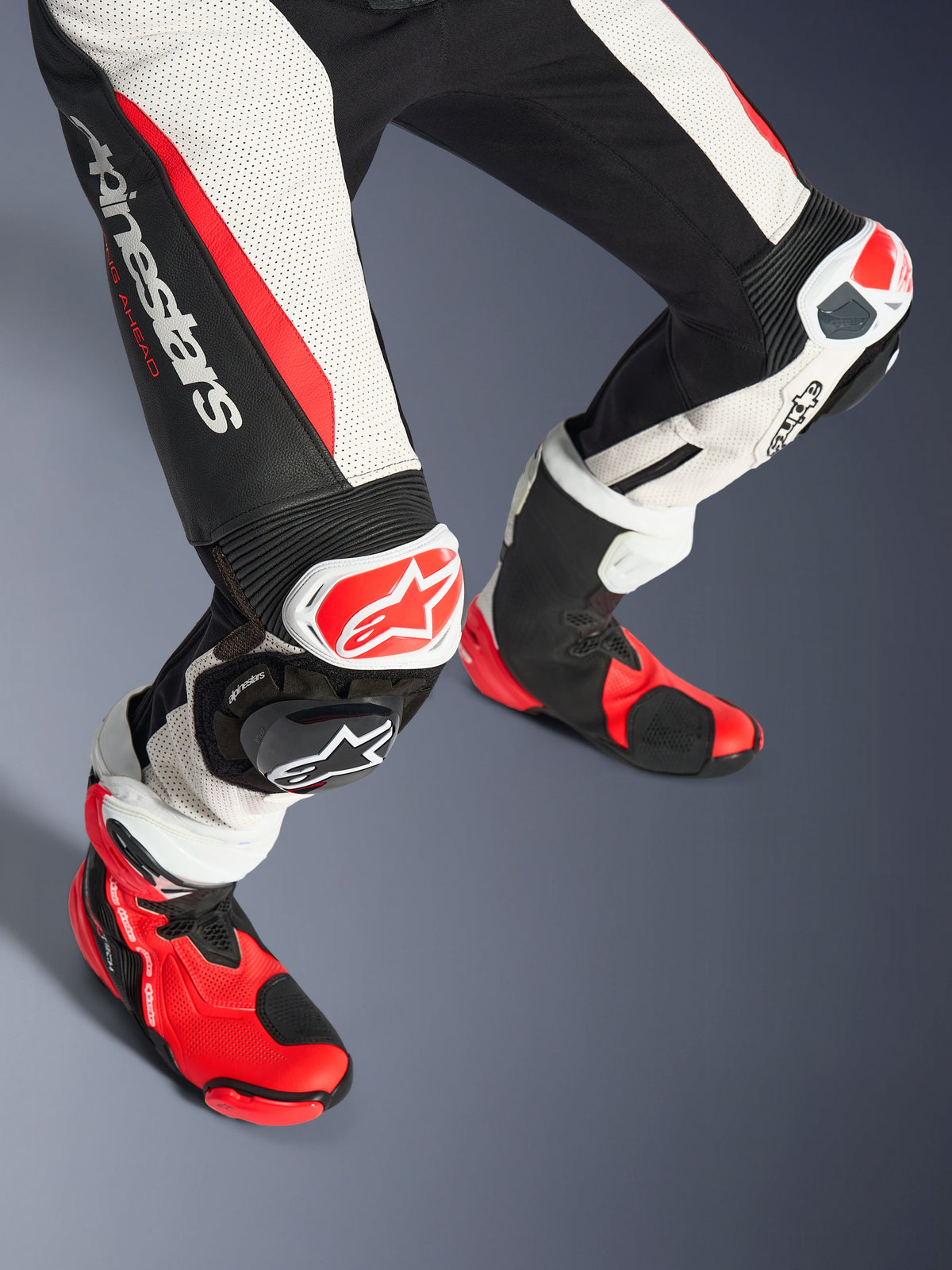 Alpinestars Stella GP-R7, dwuczęściowy damski kombinezon skórzany, dolna część nóg i buty wyścigowe, kolor czarno-biało-czerwony fluo, perforowana skóra, duże logo Alpinestars na udzie, panele harmonijkowe, czerwono-białe slidery kolan oraz dopasowane czerwono-czarne buty techniczne.