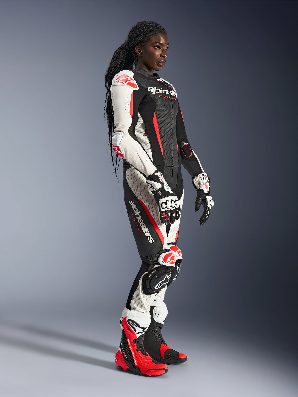 Motocyklistka w dwuczęściowym skórzanym kombinezonie Alpinestars Stella GP-R7 w kolorach czarnym, białym i czerwonym fluo, z dopasowanymi rękawicami i butami wyścigowymi, wyposażona w perforowane panele skórzane oraz ochraniacze na ramionach, łokciach i kolanach, ujęcie w studio.