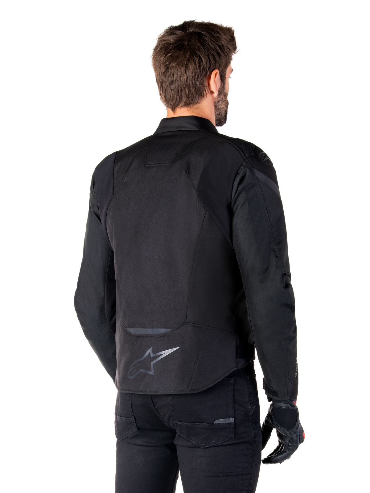Kurtka wodoodporna Alpinestars T-Jaws V4, czarna, widok z tyłu na modelu, odblaskowe logo Alpinestars w dolnej części pleców, przedłużony tył dla lepszej osłony, sportowy tekstylny design z tonalnymi panelami, noszona z czarnymi rękawicami motocyklowymi
