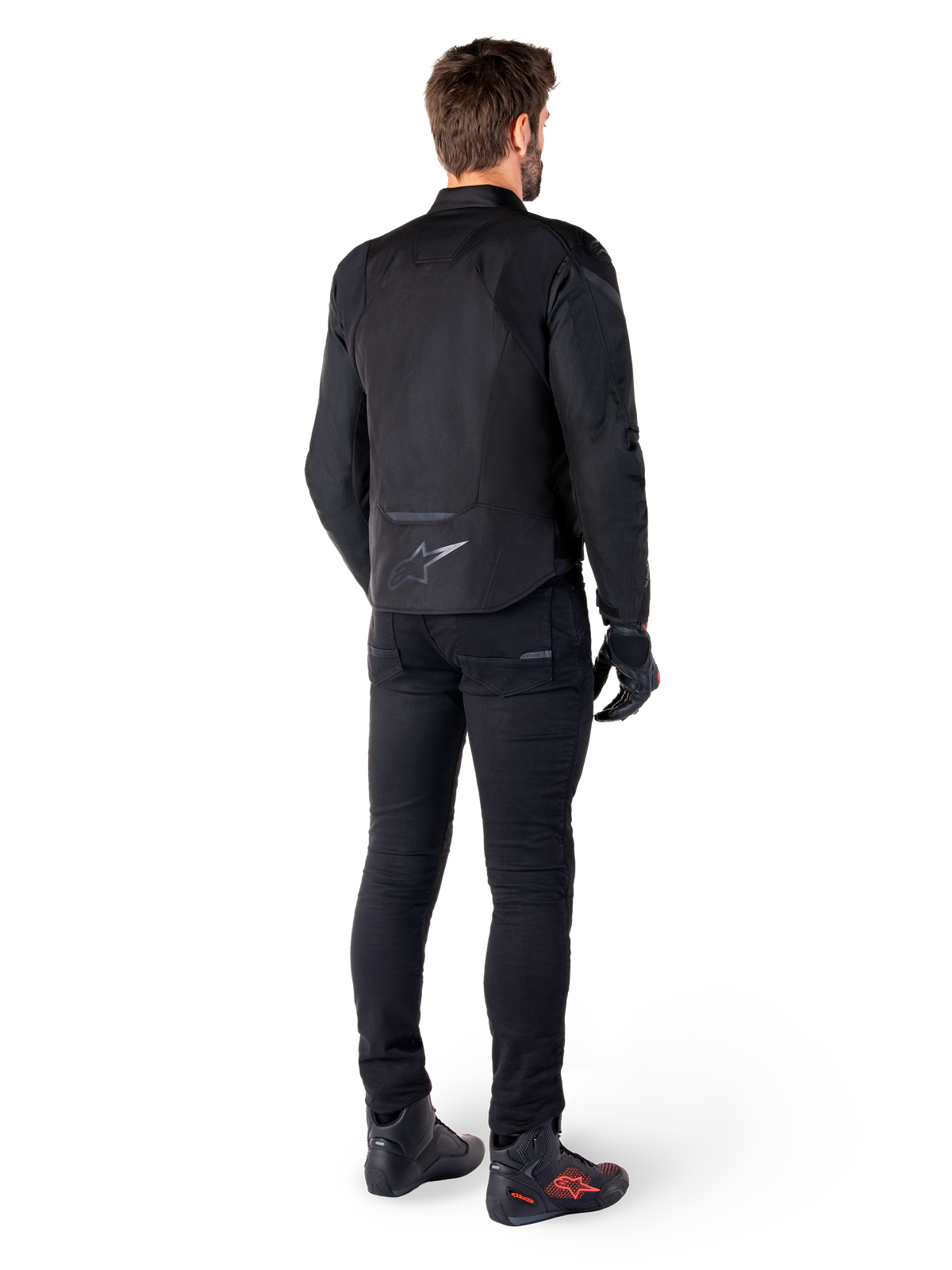 Alpinestars T-Jaws V4 Waterproof Jacket, czarna, widok z tyłu na modelu w czarnych spodniach i butach motocyklowych, sportowa konstrukcja tekstylna, logo Alpinestars w dolnej części pleców
