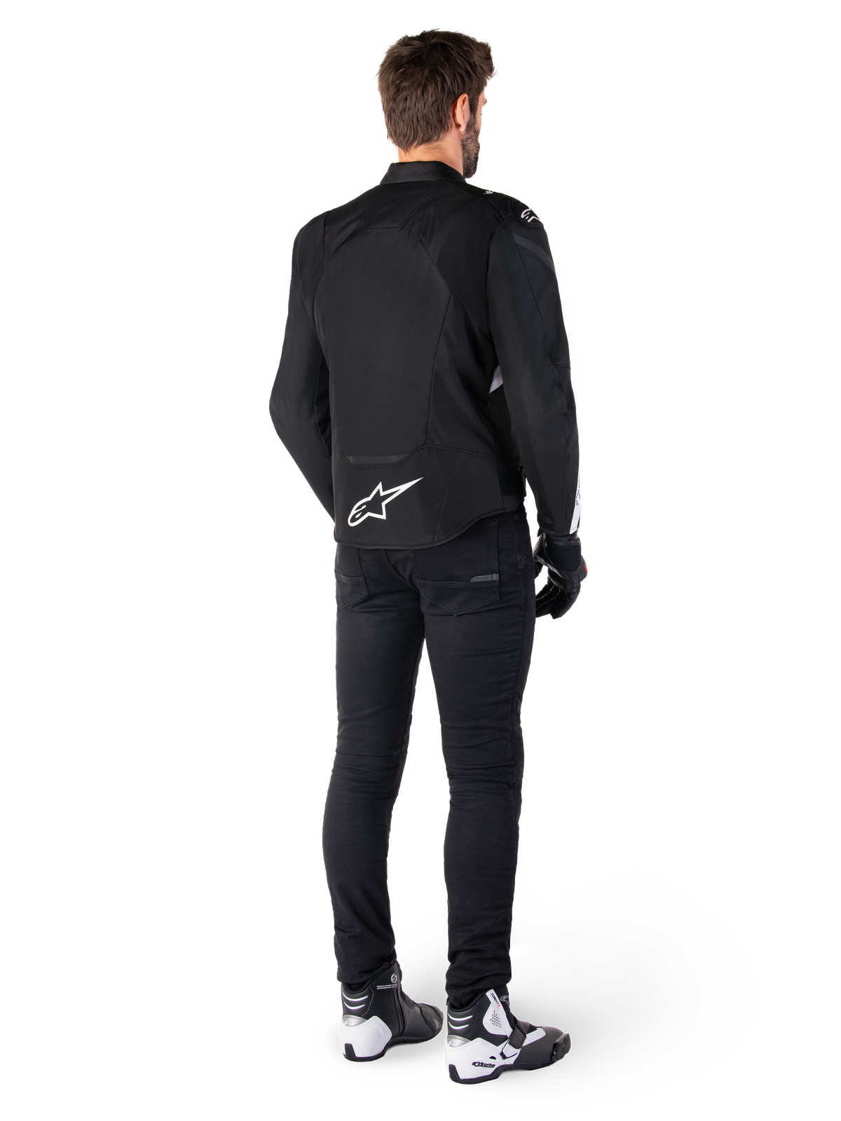 Kurtka Alpinestars T-Jaws V4 Waterproof, czarno-biała, widok z tyłu z białym logo gwiazdy w dolnej części pleców, prezentowana przez modela w czarnych spodniach motocyklowych, czarnych rękawicach oraz biało-czarnych butach motocyklowych.