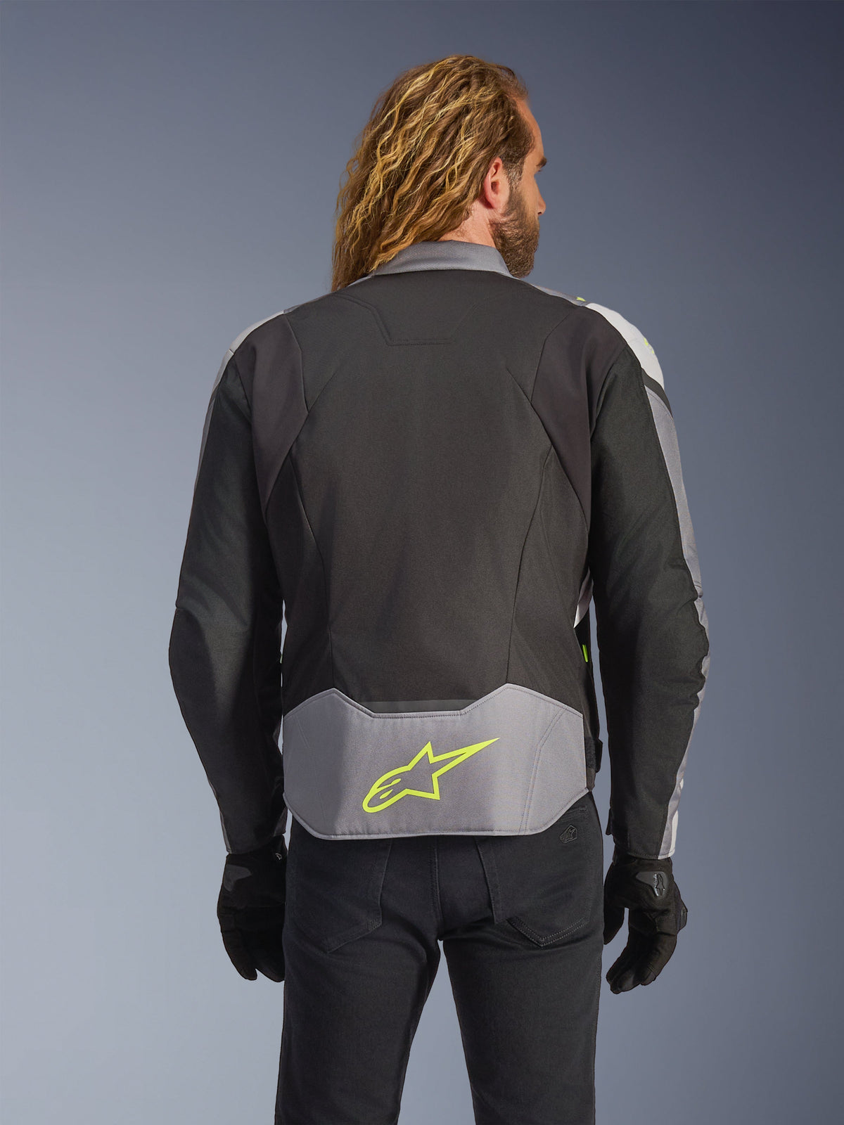 Wodoodporna kurtka Alpinestars T-Jaws V4, Black Ice Gray Dark Gray, widok z tyłu na modelu, jasnoszary panel w dolnej części pleców z wyraźnym neonowożółtym logo gwiazdy, wielopanelowa konstrukcja tekstylna, sportowa sylwetka, czarne rękawice i ciemne spodnie