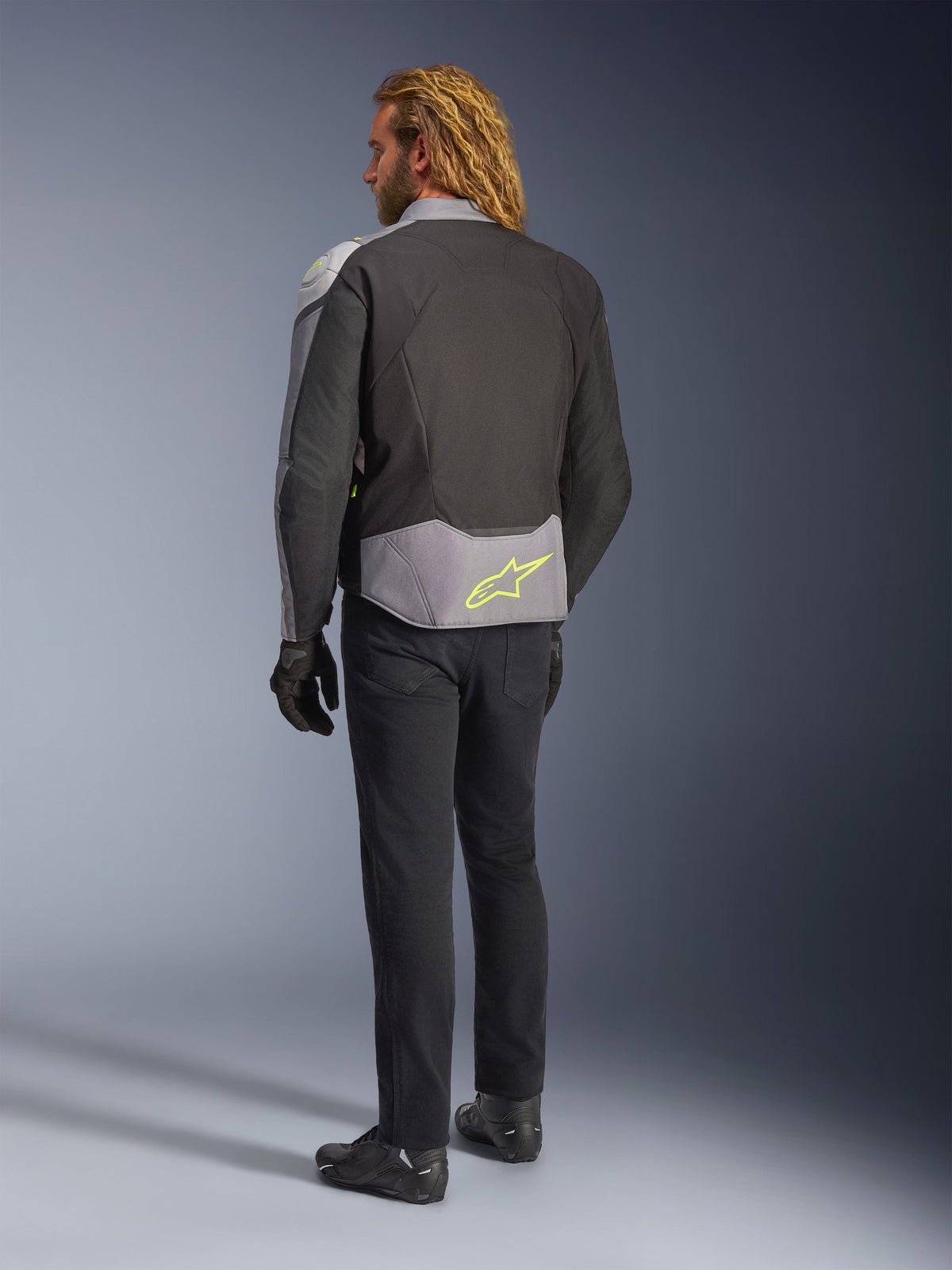 Alpinestars T-Jaws V4 Waterproof Jacket, widok z tyłu, kolorystyka Black Ice Gray i Dark Gray, z neonowożółtym logo Alpinestars w dolnej części pleców, noszona z czarnymi spodniami i rękawicami motocyklowymi, sportowa odzież motocyklowa