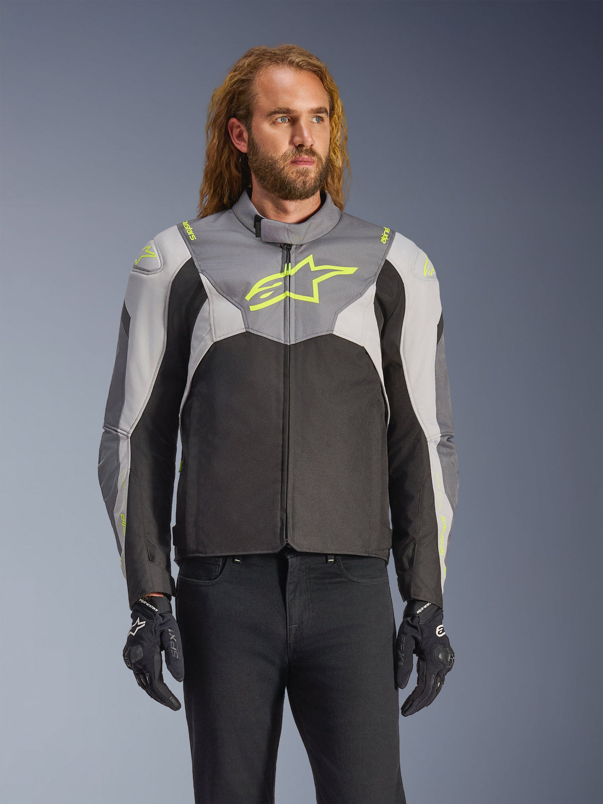 Alpinestars T-Jaws V4 Waterproof Jacket noszona przez modela, kolory Black Ice Gray i Dark Gray z neonowożółtymi akcentami, z wyraźnym żółtym logo gwiazdy na klatce piersiowej, stylizowana z czarnymi spodniami i rękawicami motocyklowymi, tekstylna kurtka motocyklowa o sportowym kroju
