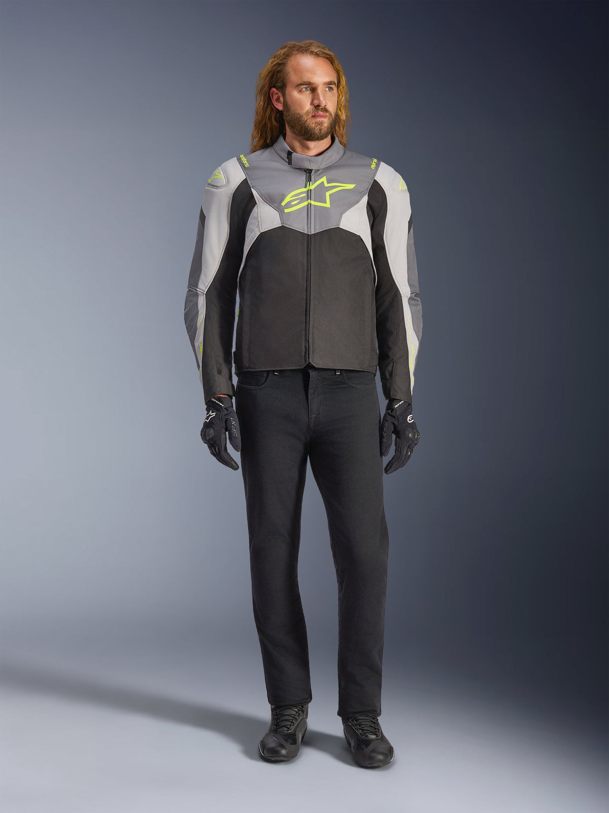 Alpinestars T-Jaws V4 Waterproof Jacket, wodoodporna kurtka motocyklowa, czarna, lodowo-szara i ciemnoszara z neonowożółtymi akcentami, prezentowana przez modela, sportowa sylwetka, techniczna konstrukcja tekstylna, wyraźne logo na piersi, zestawiona z czarnymi spodniami i rękawicami