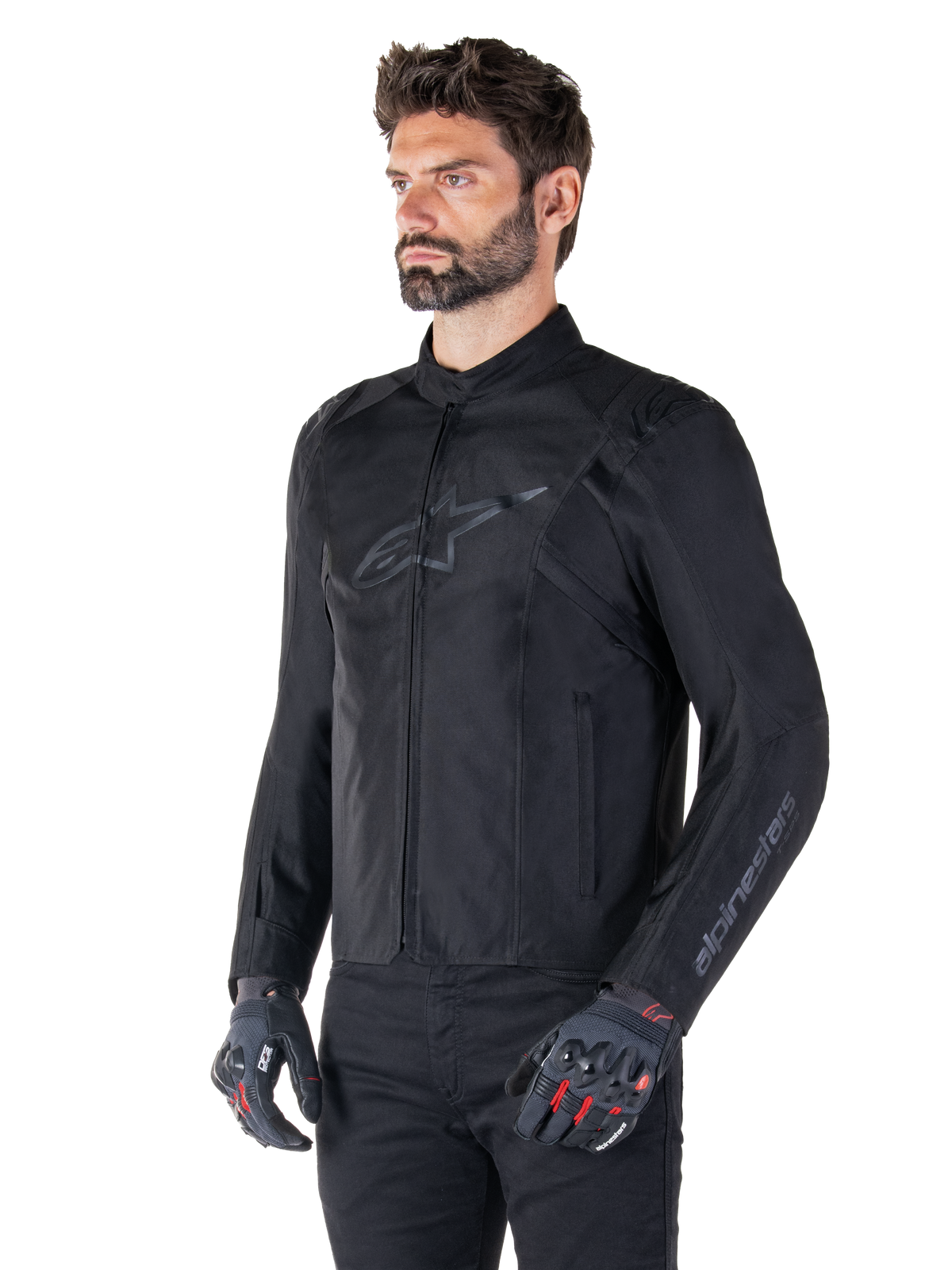 Model w kurtce Alpinestars T-SPS V2 Waterproof Jacket, czarna, zestawiona z czarnymi spodniami i szarymi rękawicami motocyklowymi z czerwonymi akcentami, subtelne czarne logo na piersi i branding na rękawach, tekstylna kurtka o sportowym kroju do jazdy szosowej.