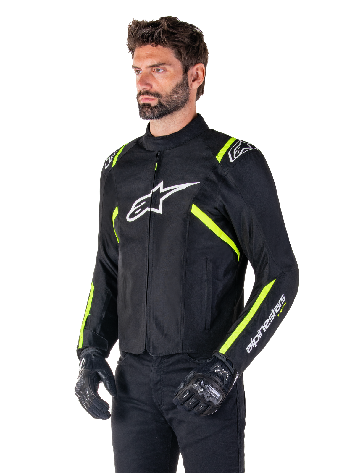 Męski model w kurtce Alpinestars T-SPS V2 Waterproof, czarnej z białymi i żółtymi neonowymi akcentami, z białym logo Astars na piersi i neonowożółtymi paskami na ramionach i rękawach, w zestawie z czarnymi spodniami i rękawicami motocyklowymi, tekstylna kurtka motocyklowa o sportowym kroju