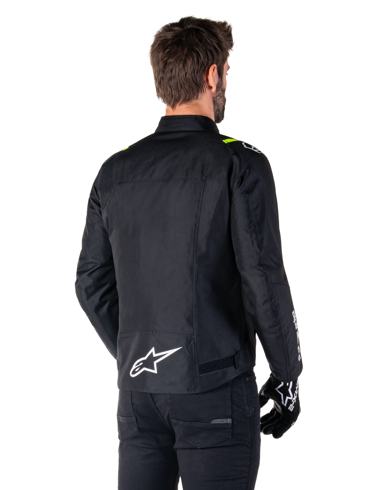 Alpinestars T-SPS V2 Waterproof Jacket, czarna z białymi i żółtymi akcentami fluo, widok z tyłu na modela ubranego w tekstylną kurtkę motocyklową, białe logo w dolnej części pleców, żółte akcenty fluo na ramionach, czarne spodnie i rękawice