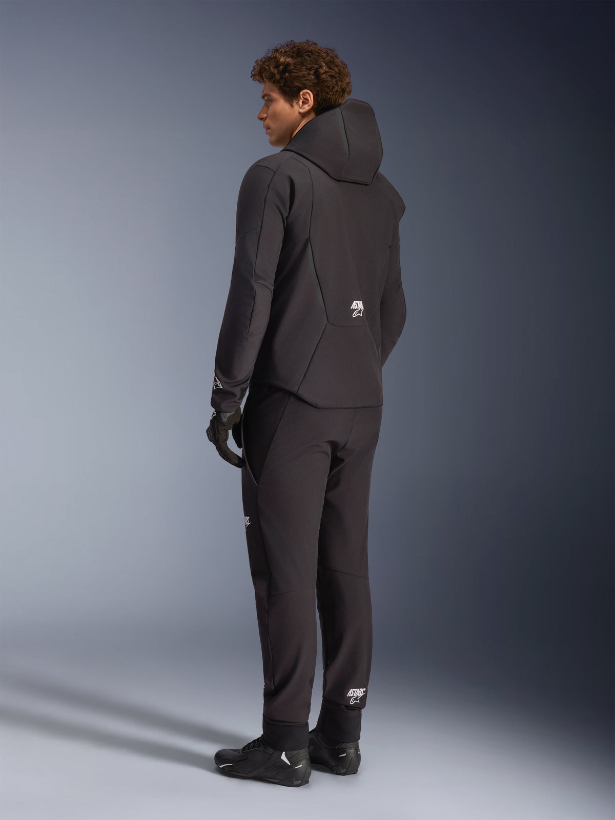 Alpinestars Aeroshell Waterproof Jacket, męska kurtka motocyklowa z kapturem, czarny tekstylny softshell, wodoodporna, widok z tyłu na modelu, białe logo Alpinestars, odzież szosowa i touring