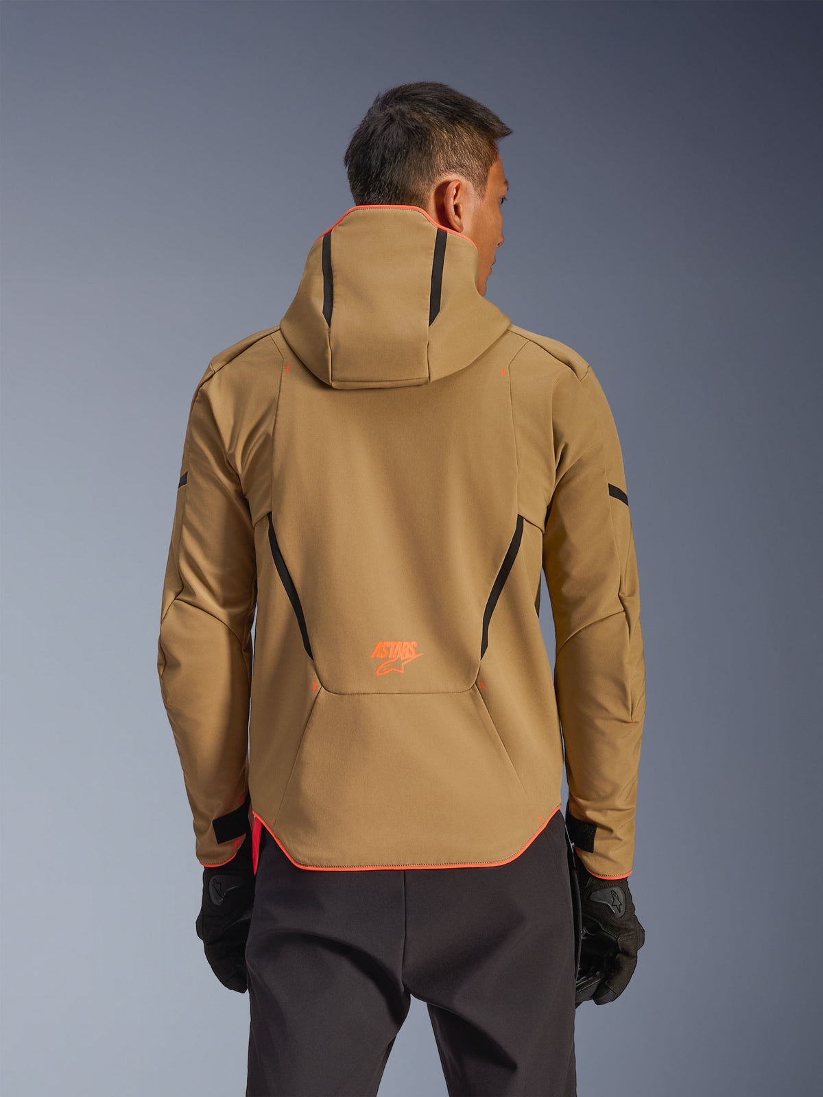 Wodoodporna kurtka Alpinestars Aeroshell, Dark Khaki i Orange Fluo, widok z tyłu prezentujący fason bluzy z kapturem z pomarańczowym wykończeniem kaptura i dolnej krawędzi, pomarańczowe logo w dolnej części pleców, zestawiona z czarnymi rękawicami, sportowa odzież motocyklowa typu softshell crossover.