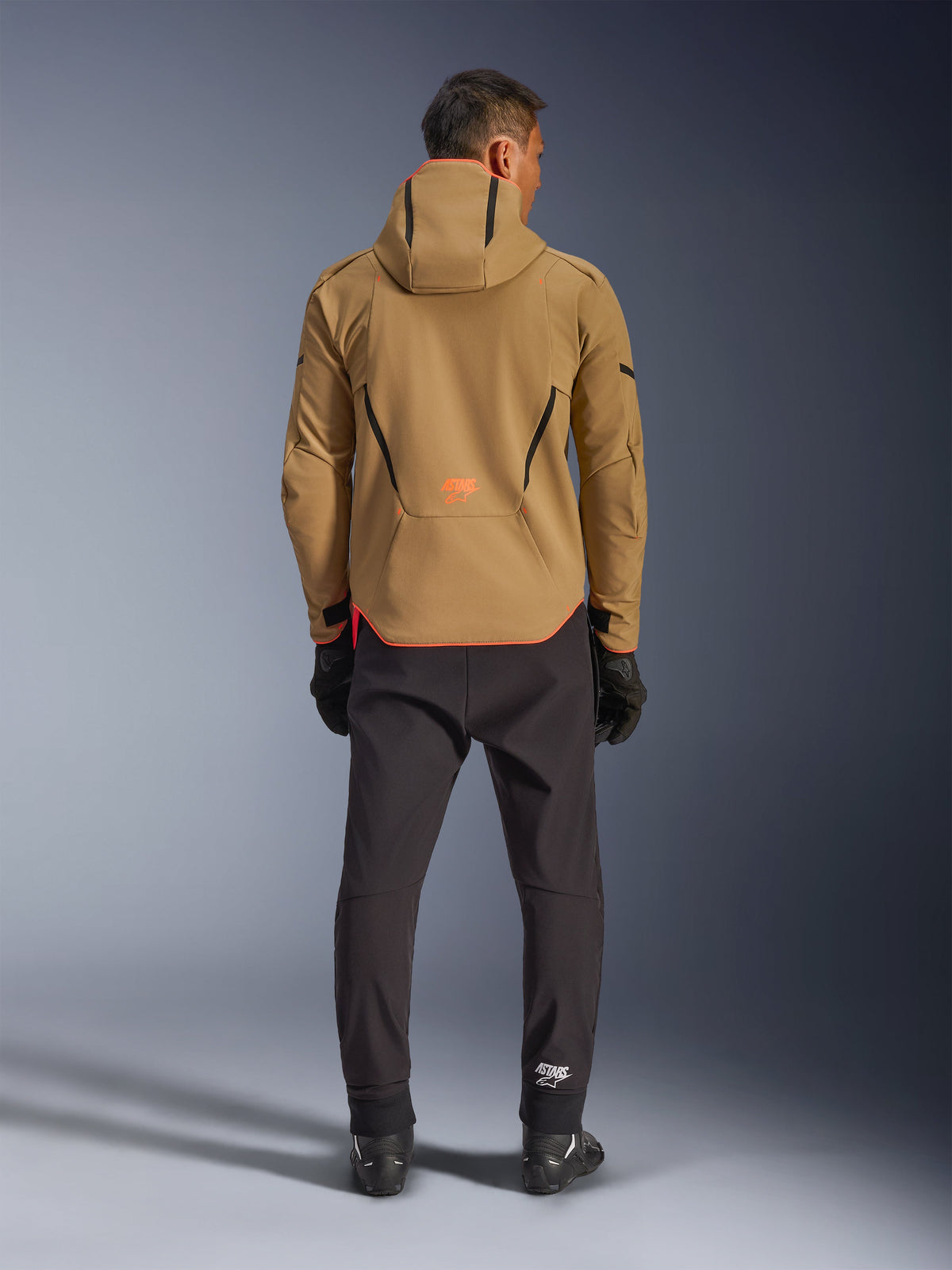 Wodoodporna kurtka Alpinestars Aeroshell, kolor Dark Khaki Black Orange Fluo, widok od tyłu na mężczyznę w kurtce softshell z kapturem, czarne lamówki i pomarańczowe logo, czarne spodnie techniczne i rękawice