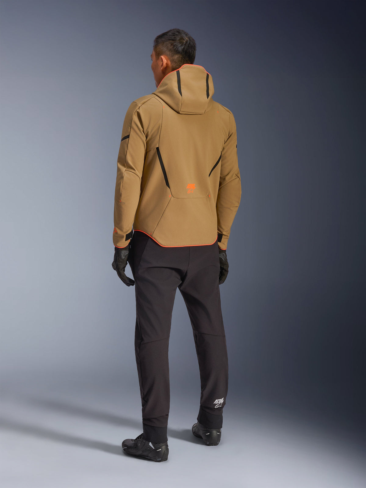 Alpinestars Aeroshell Waterproof Jacket, Dark Khaki Black Orange Fluo, widok od tyłu motocyklisty w kurtce softshell z kapturem, czarne spodnie i rękawice, czarne geometryczne akcenty i pomarańczowe logo na dole pleców