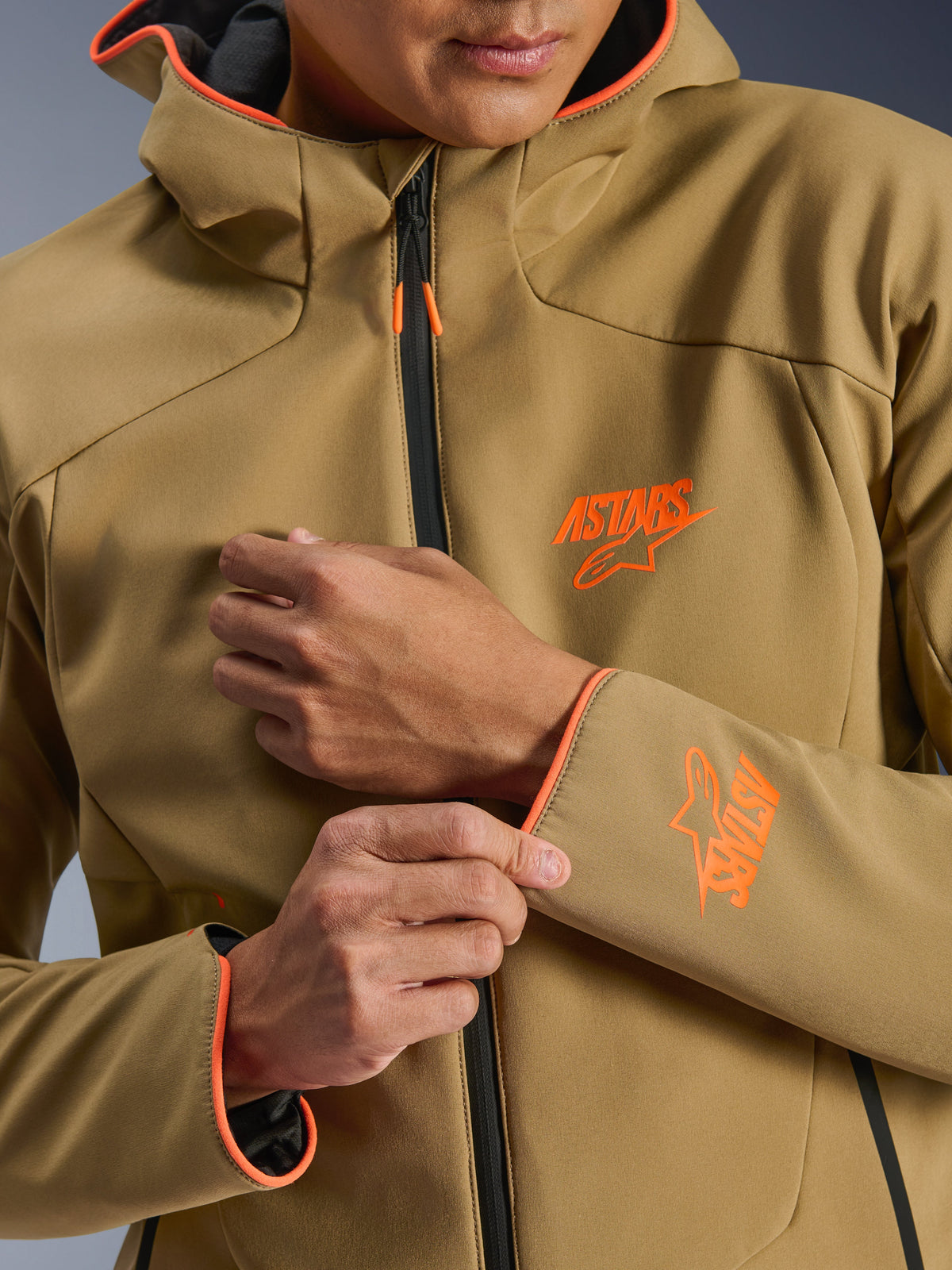 Alpinestars Aeroshell Waterproof Jacket, ciemne khaki i jaskrawy pomarańcz (Orange Fluo), zbliżenie na przód i rękaw z pomarańczowymi logo ASTARS, pomarańczowe lamówki na kapturze i mankietach, czarny wodoodporny zamek błyskawiczny.