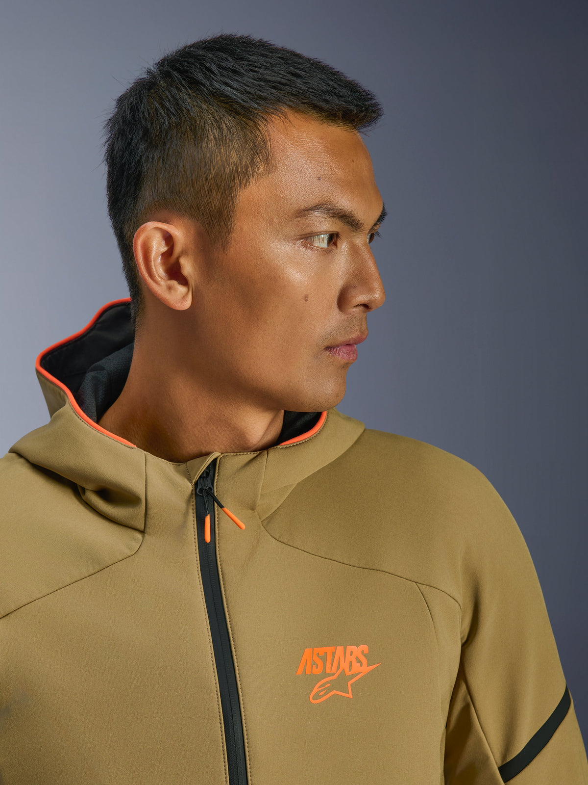 Kurtka Alpinestars Aeroshell Waterproof, kolor ciemne khaki i jaskrawy pomarańczowy, zbliżenie z profilu, pomarańczowe logo ASTARS na piersi i kaptur z pomarańczową podszewką, nowoczesna bluza motocyklowa typu softshell